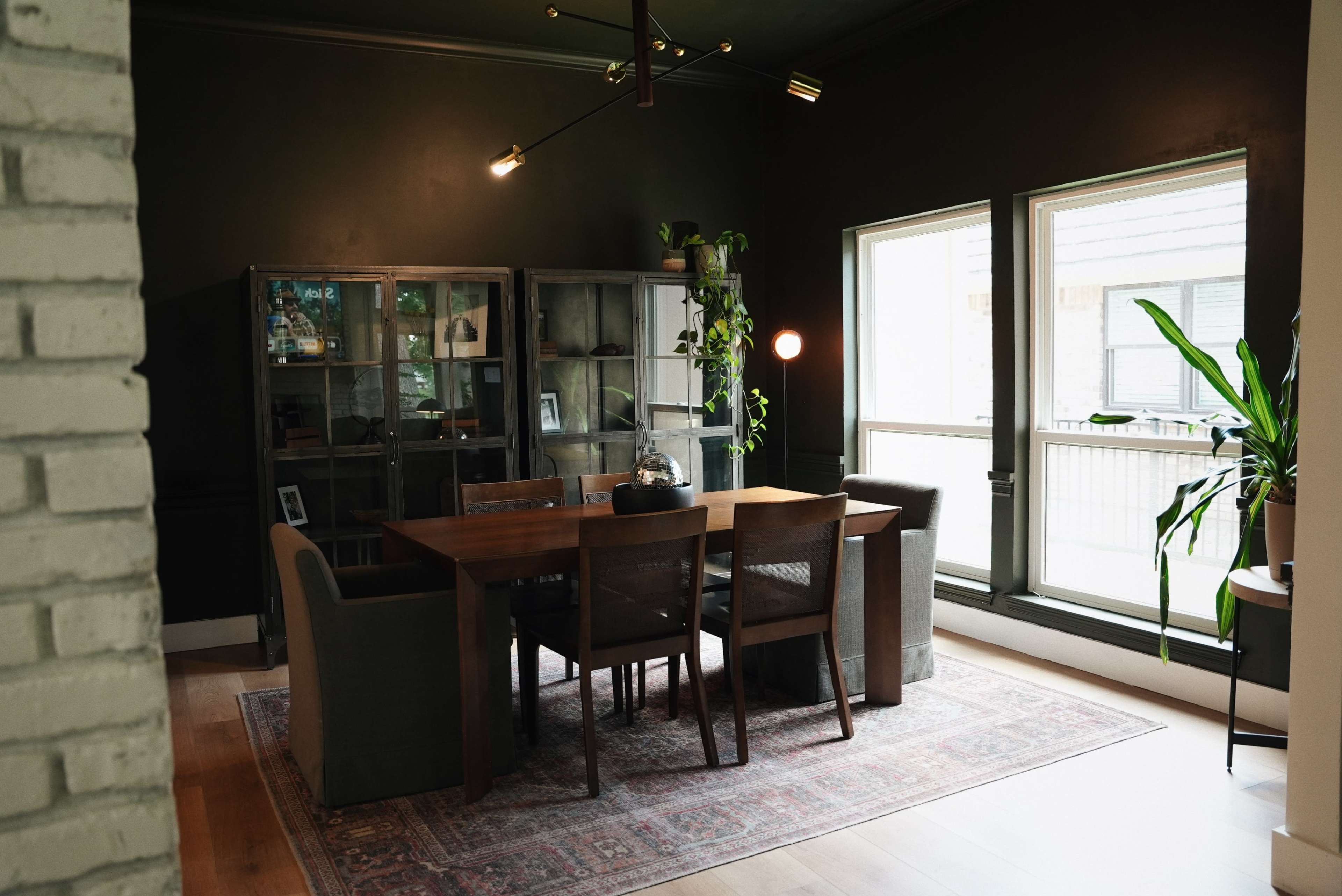The image shows a modern dining room with a wooden table surrounded by chairs, set against dark walls and featuring large windows.