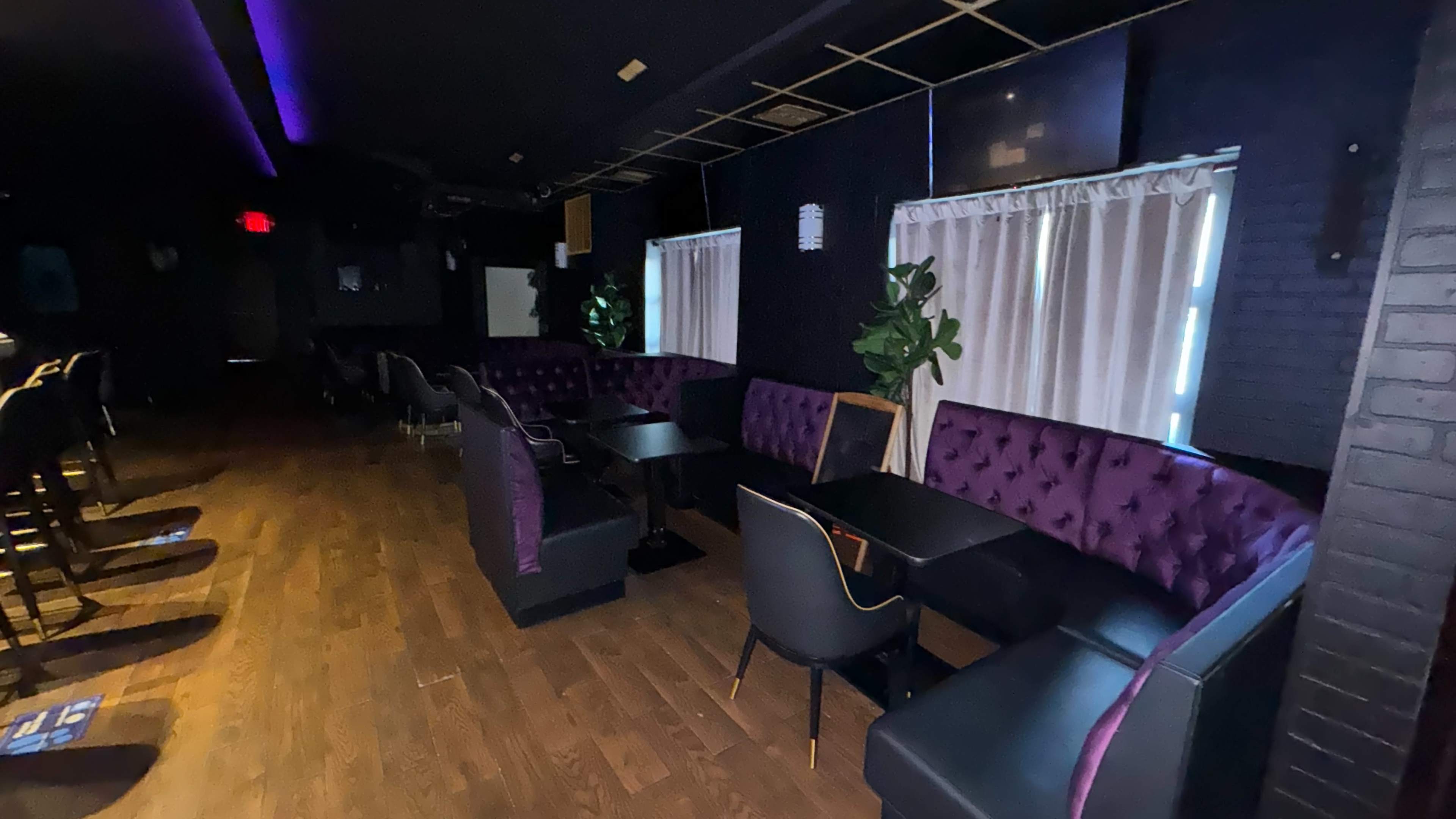 The image shows a dimly lit bar or lounge area featuring purple upholstered seating, empty tables, and large windows with sheer curtains.