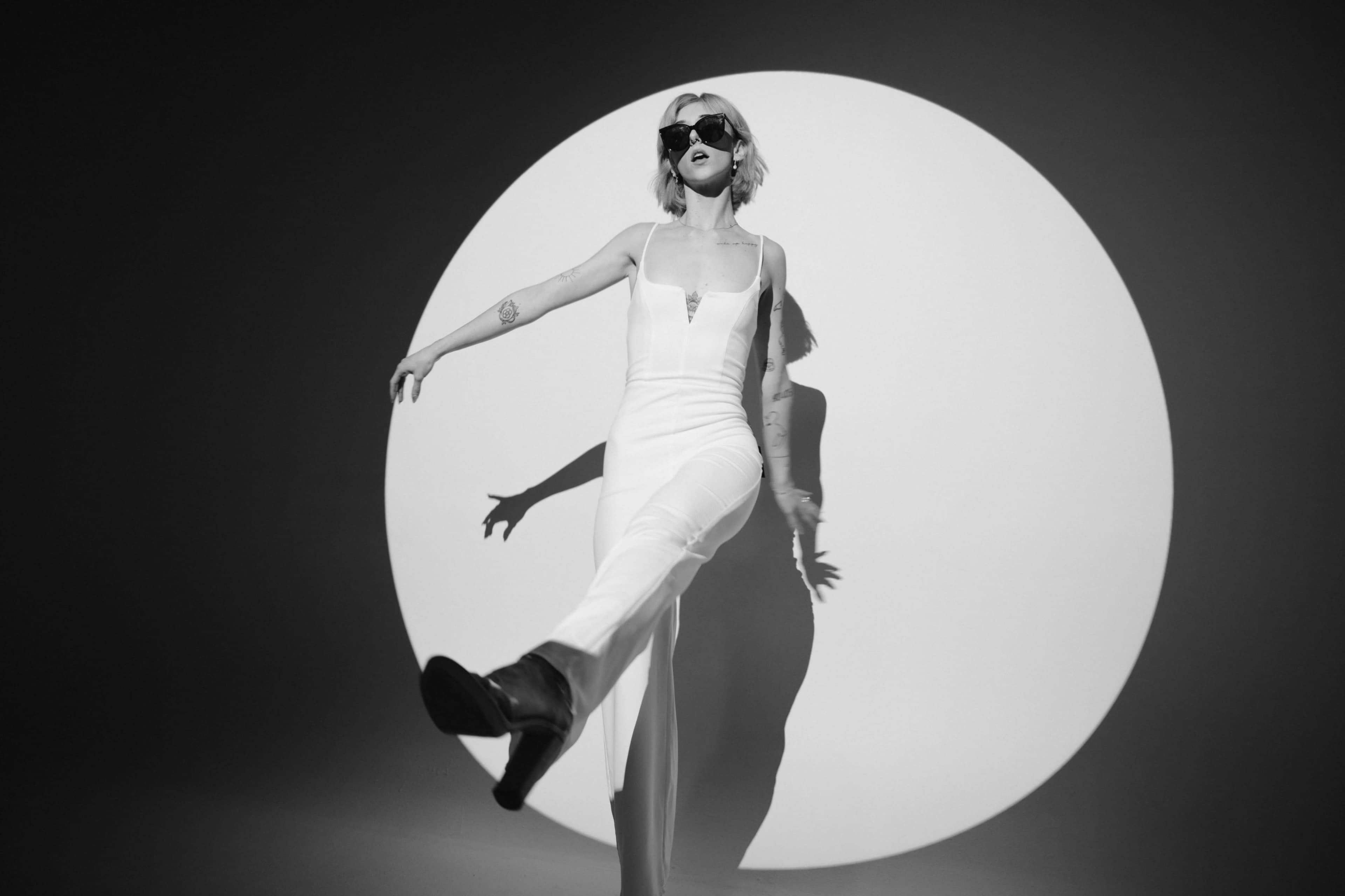 A person in a white outfit strikes a pose with one leg extended in front of a large circular backdrop.