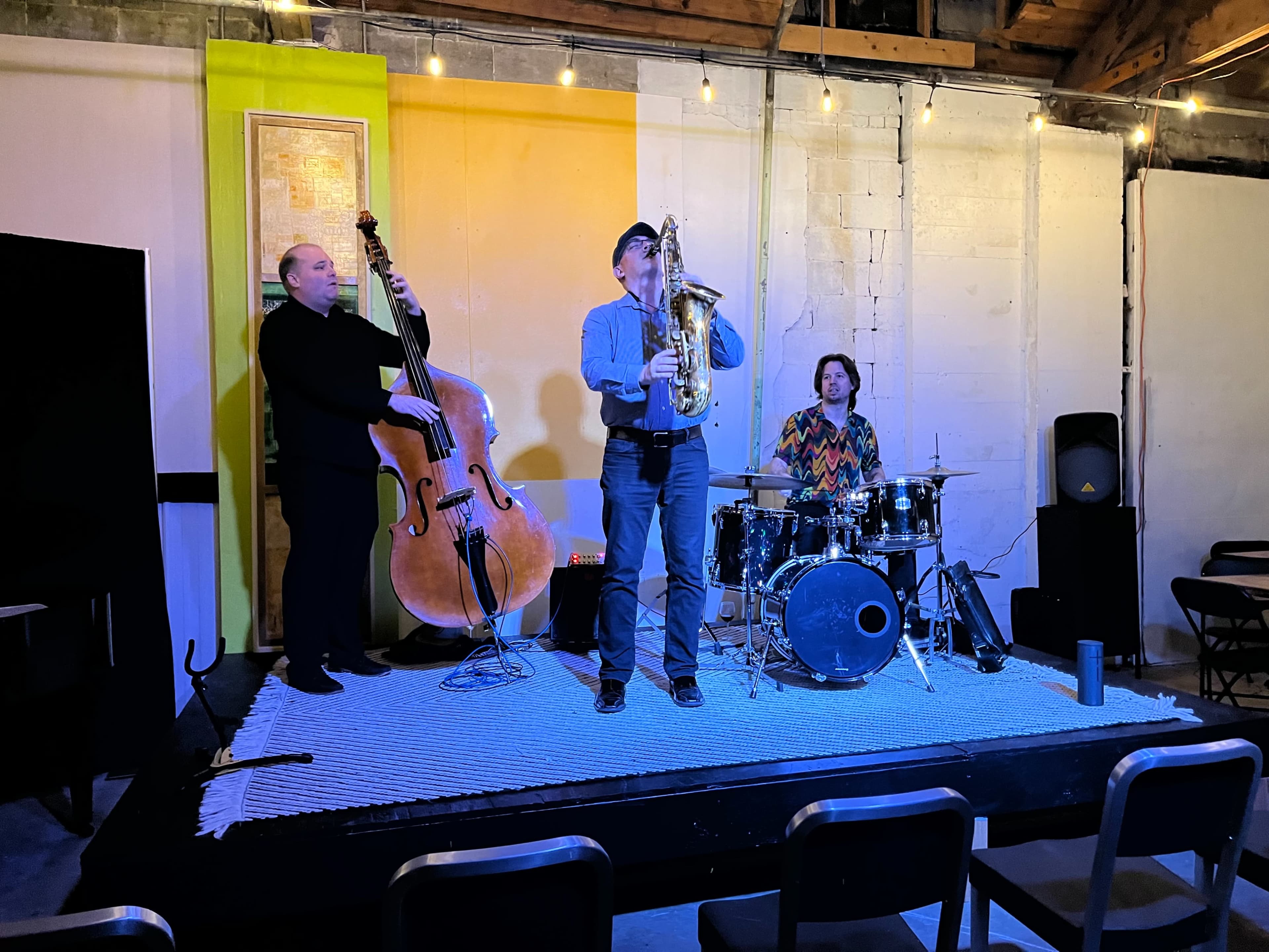 Three musicians perform on a small stage with a double bass, saxophone, and drums, in a dimly lit room with colorful wall decorations.