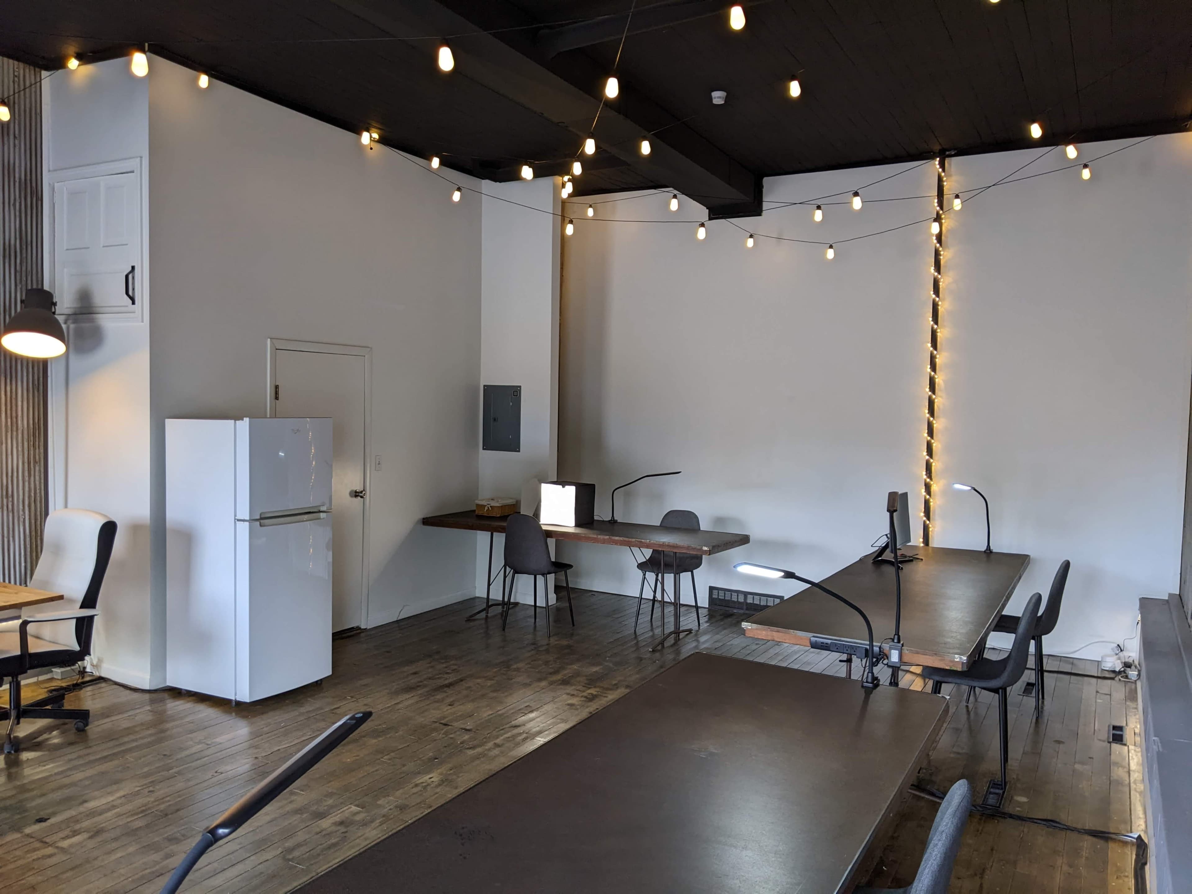 The image shows a spacious, well-lit office with several desks, chairs, a refrigerator, and string lights hanging from the ceiling.