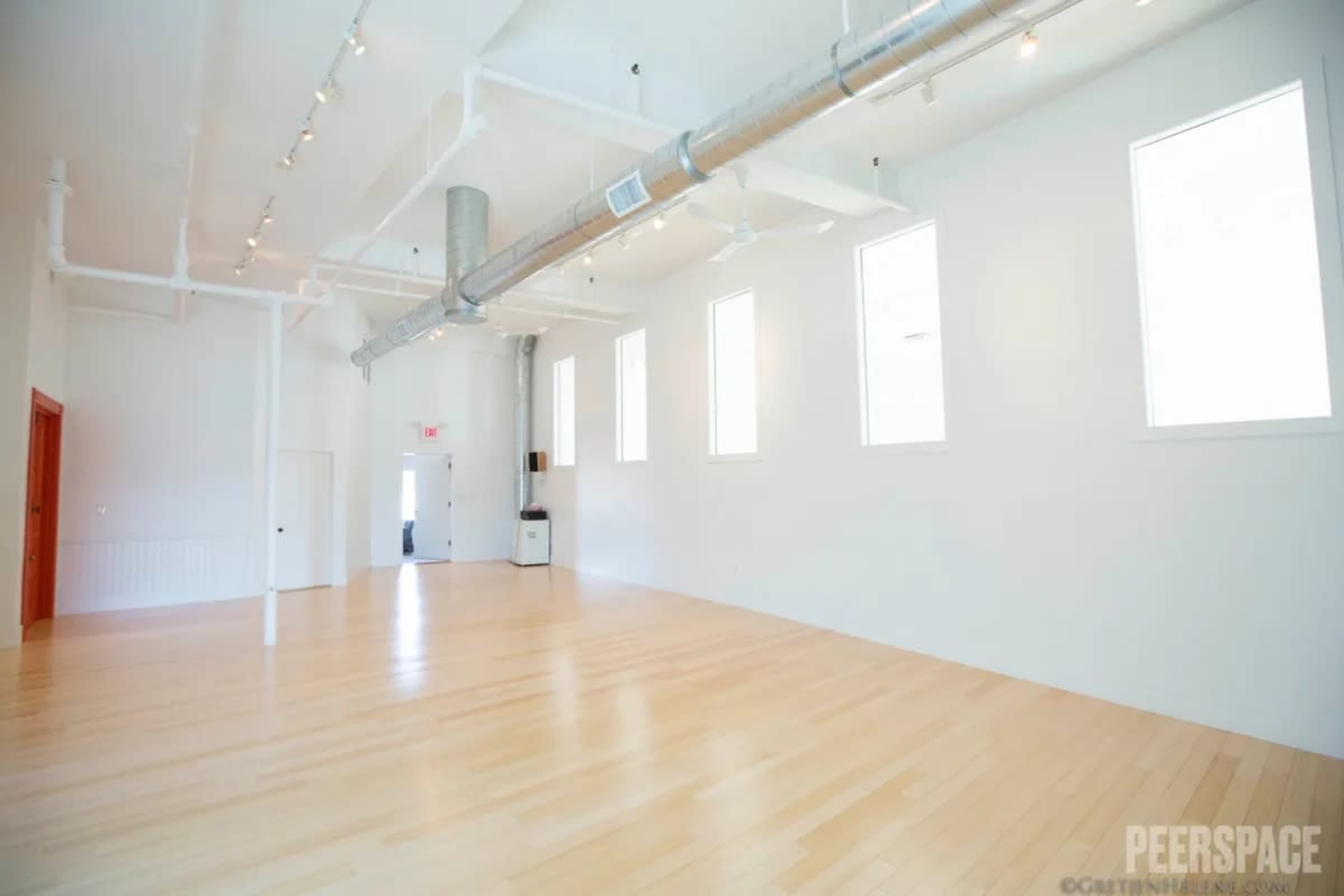 The image shows a spacious, empty room with wooden flooring, white walls, and large windows allowing natural light.