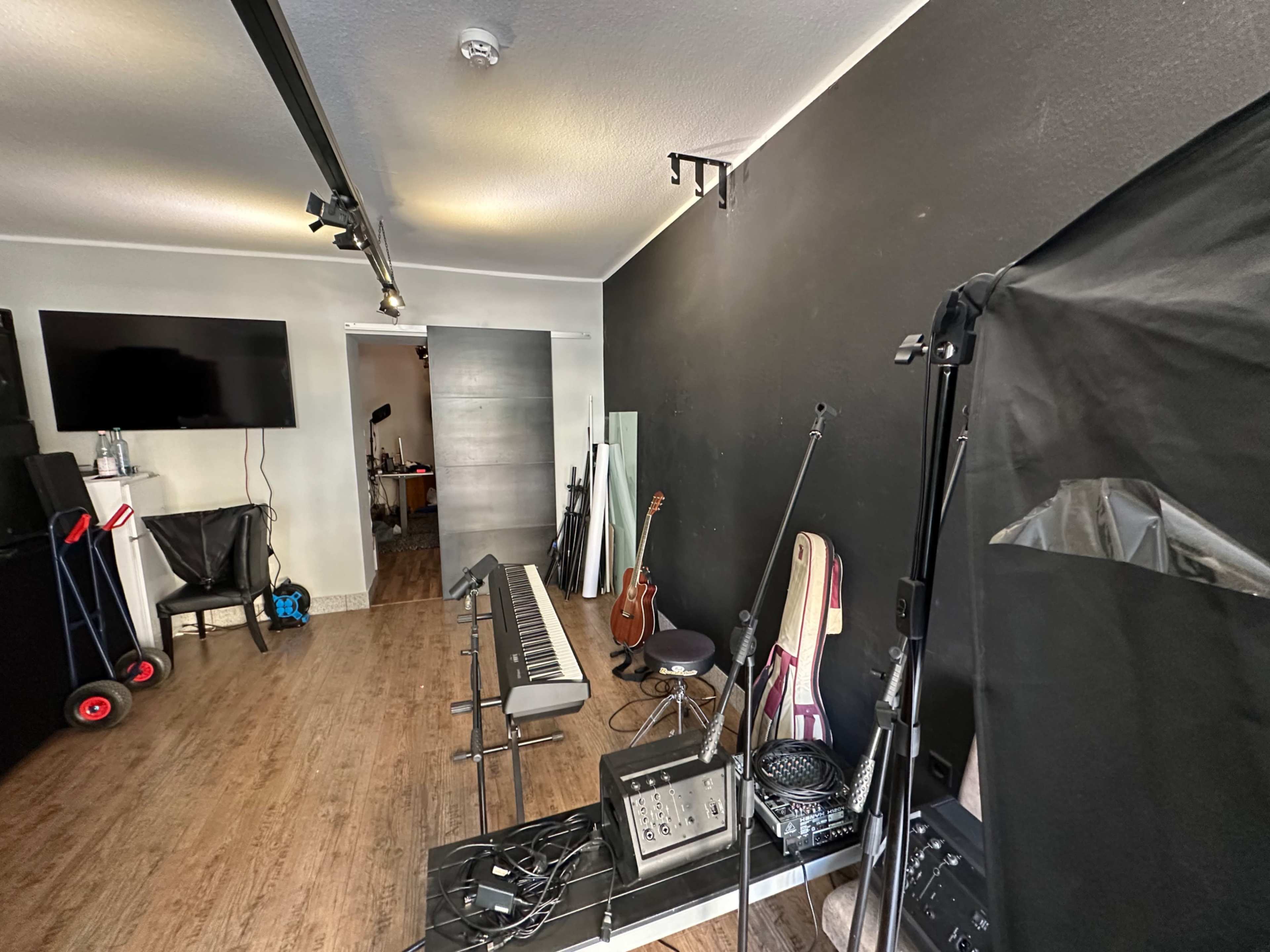 The image shows a music studio with a black wall, various instruments, and sound equipment scattered on the floor, along with a seating area and a television on one side.