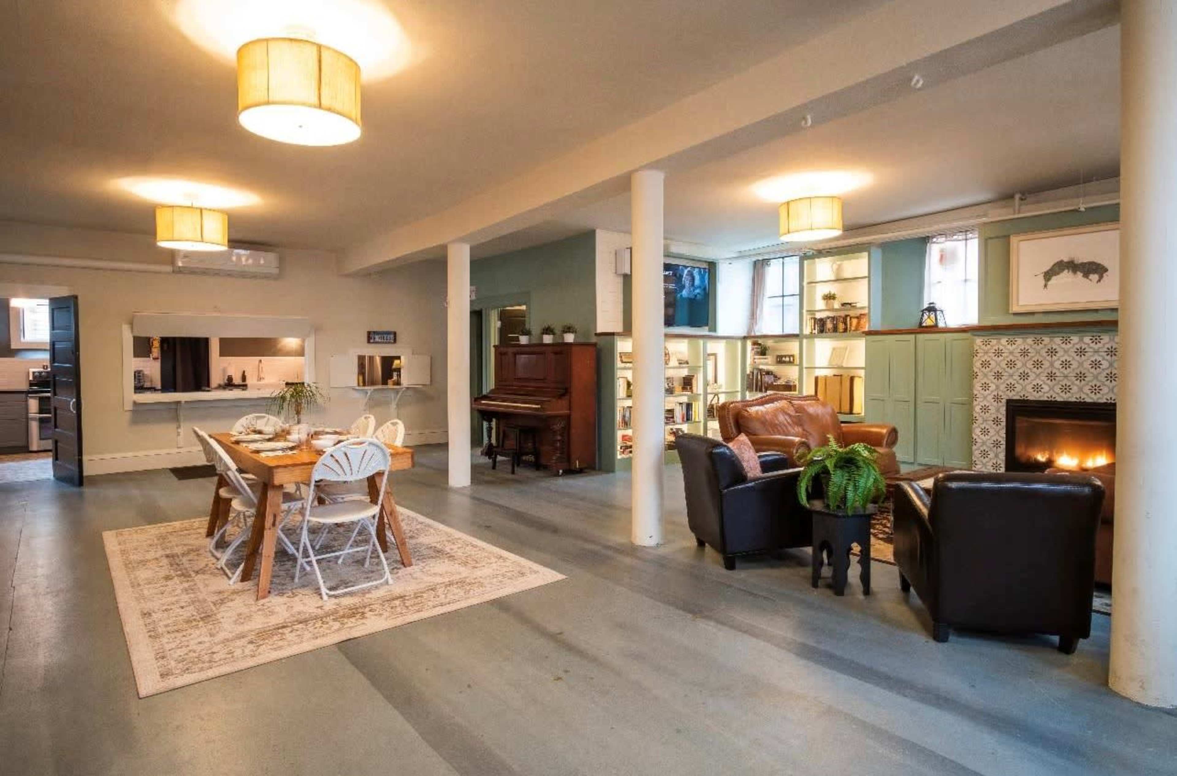 The image shows a spacious room featuring a dining area with a table and chairs, a living area with a sofa and armchairs, a piano, and a fireplace, along with decorative shelves and light fixtures.