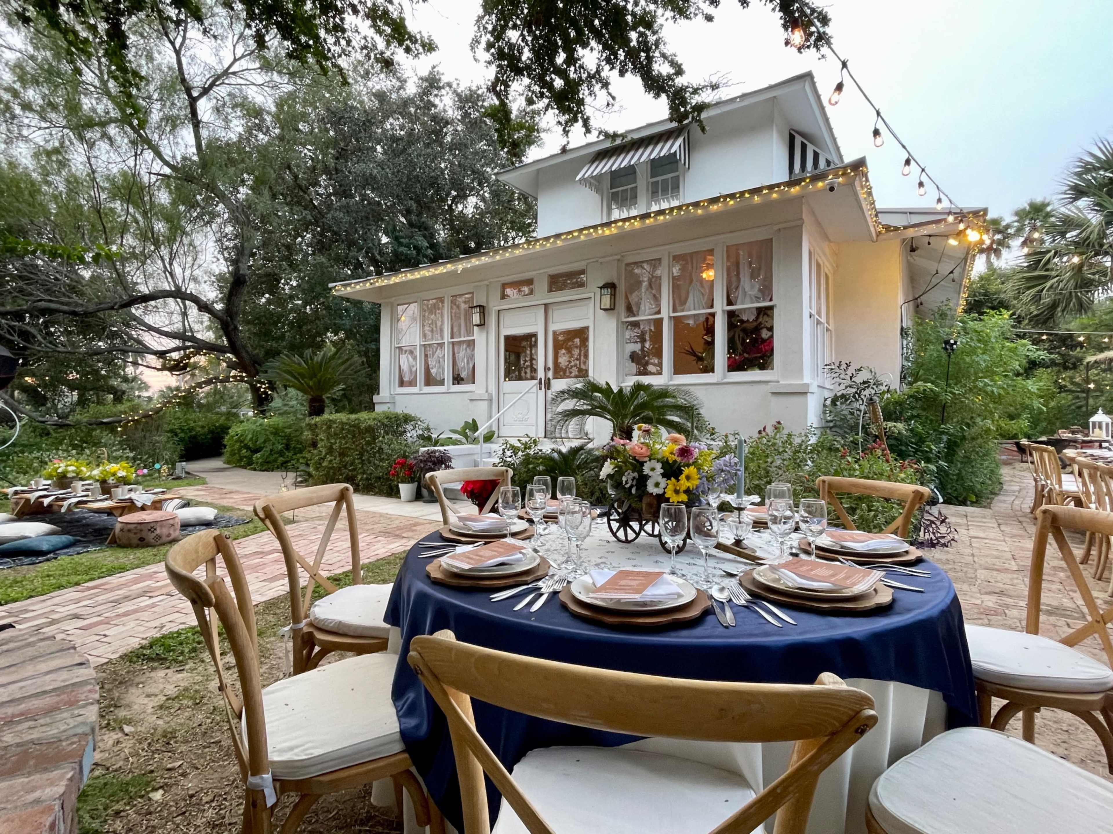A beautifully set dining table with elegant tableware is arranged in front of a charming white house surrounded by greenery and flowers.