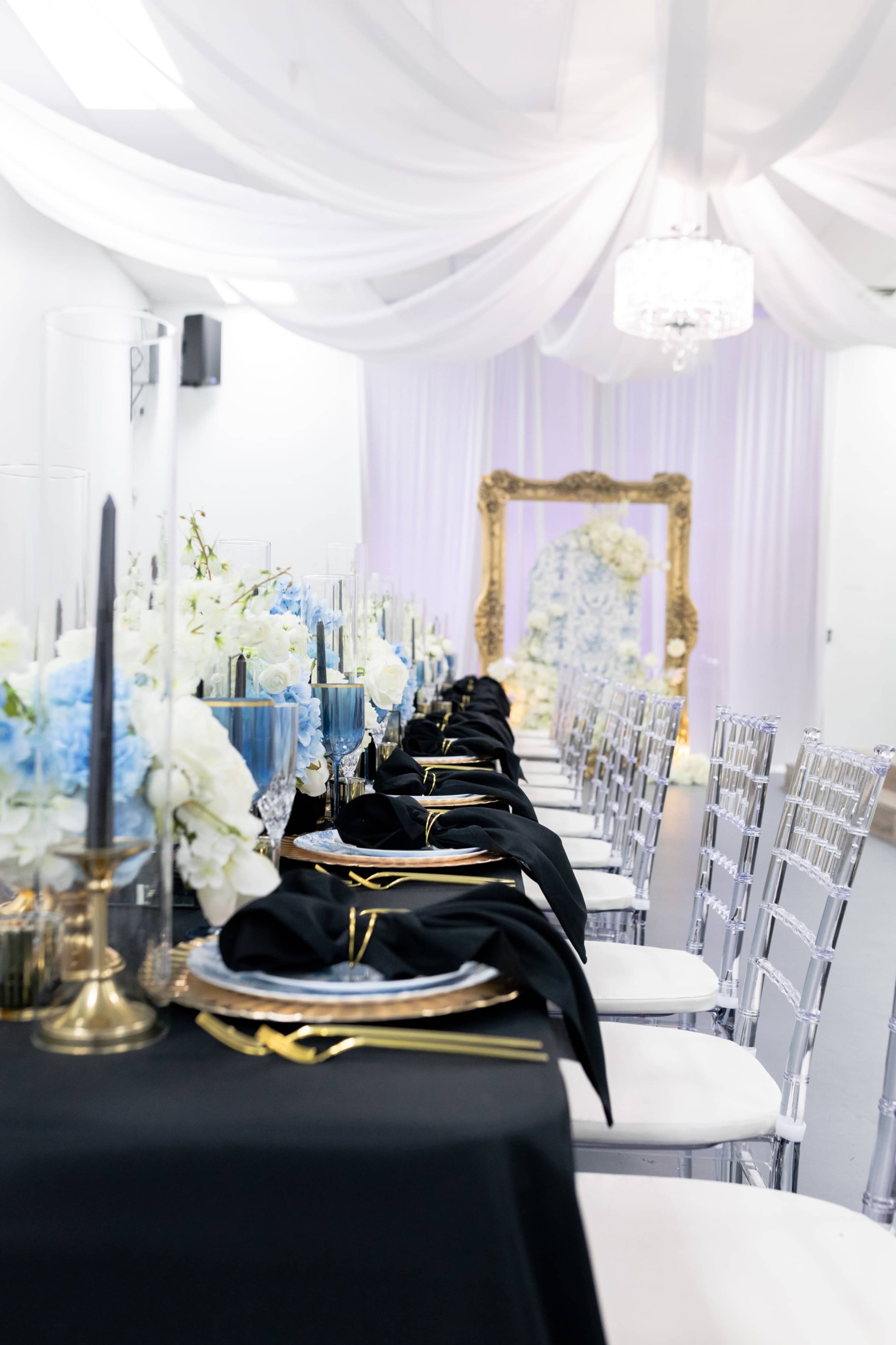 A long banquet table is set with black tablecloths, gold and silver tableware, and adorned with floral arrangements in shades of blue and white, under a draped ceiling with a chandelier.