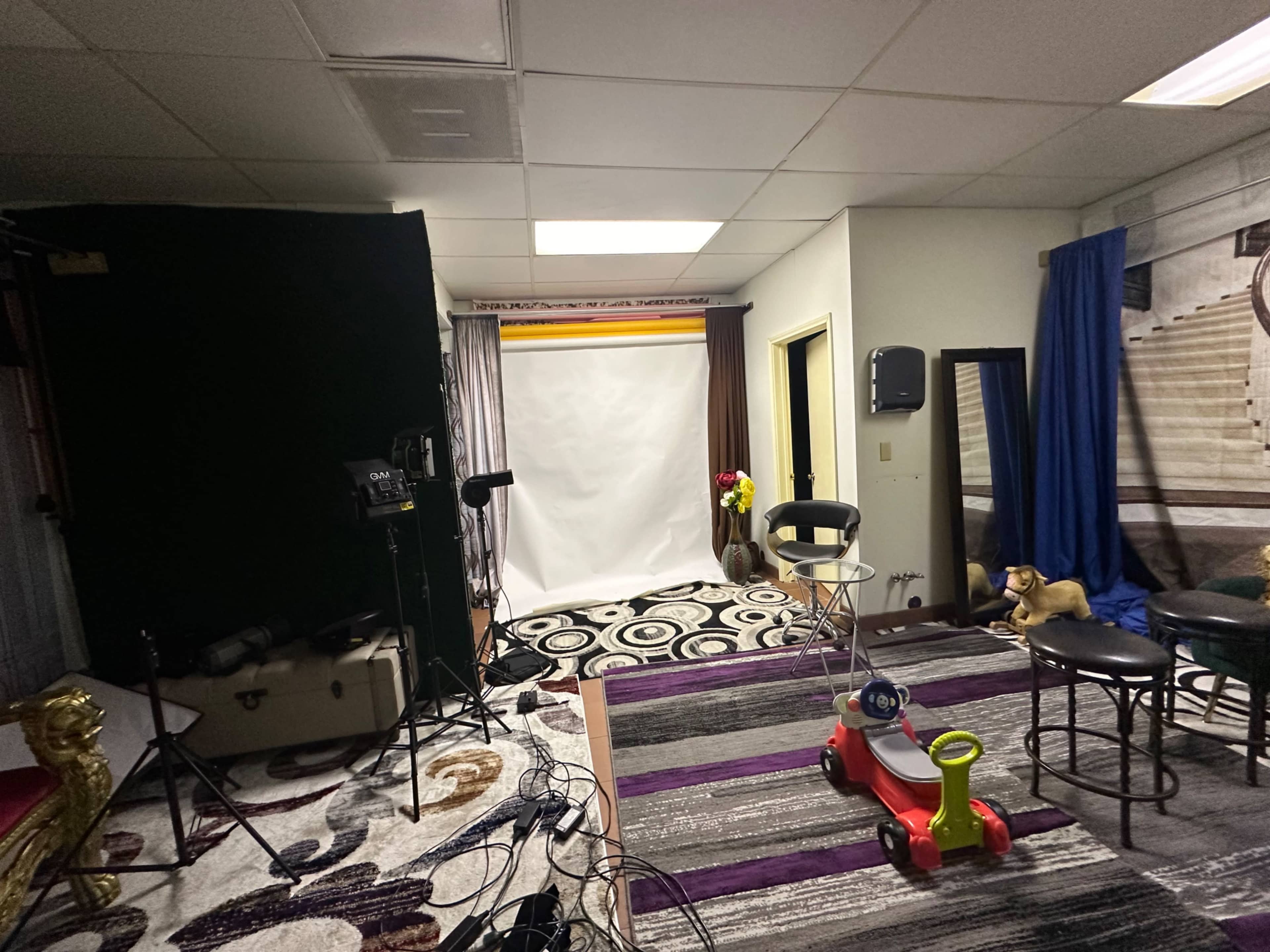 The image shows a cluttered studio space with photography equipment, a blank backdrop, a rug, and a toy car on the floor.
