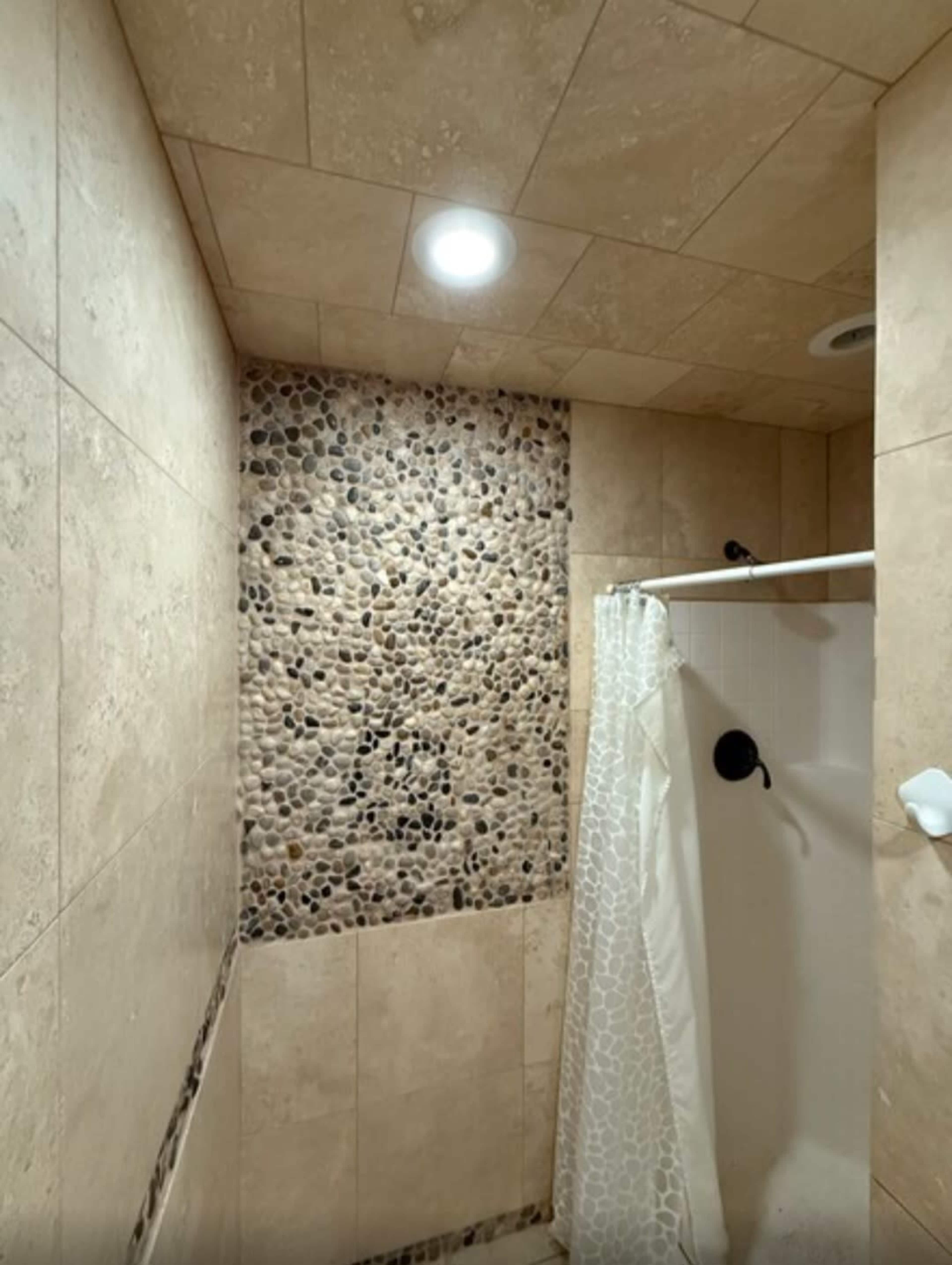 The image shows a shower area with beige tiled walls, a pebble mosaic accent wall, and a white shower curtain.