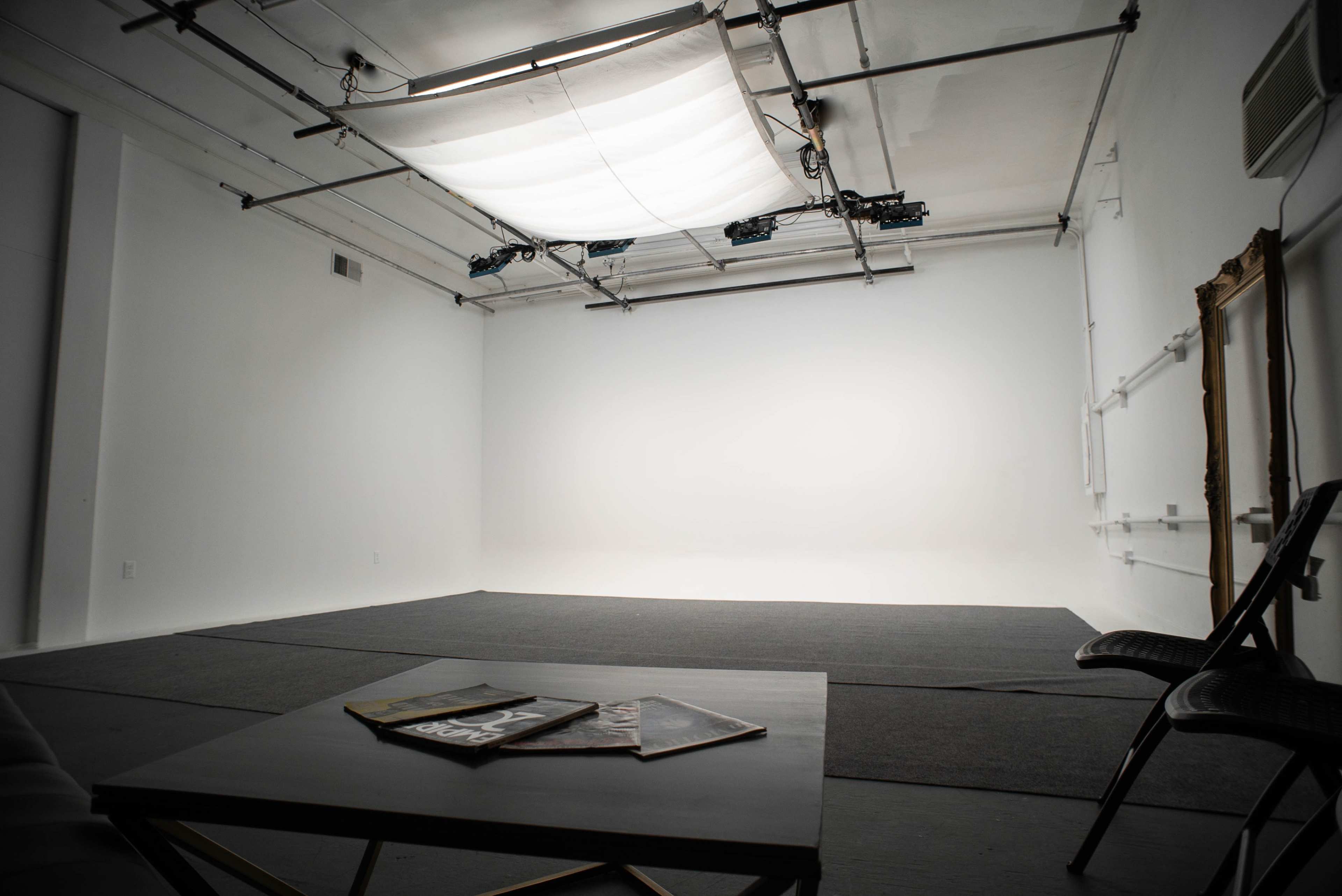 An empty, well-lit studio space with gray flooring, a coffee table displaying magazines, and two chairs positioned nearby.