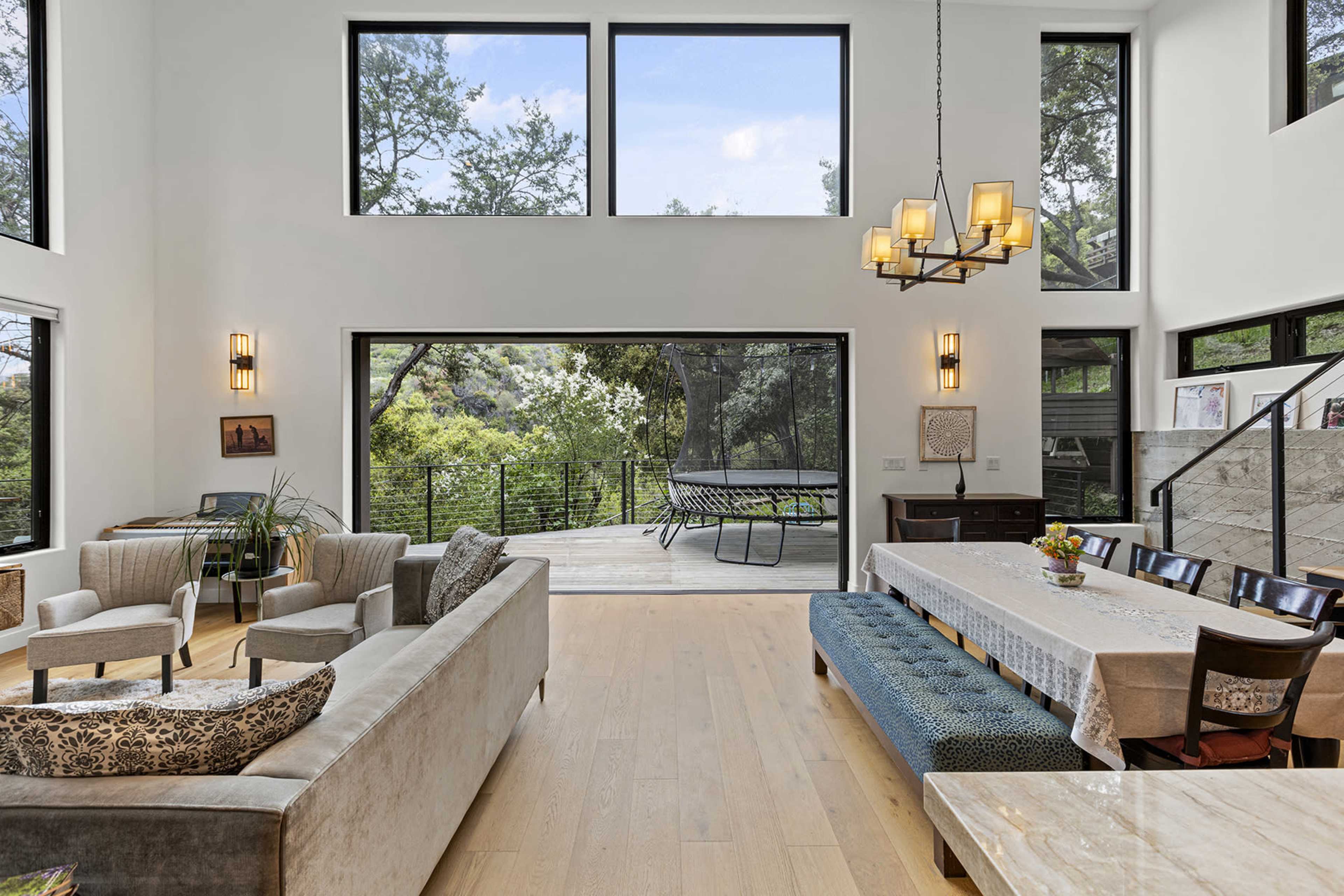 A modern interior features a spacious living room with large windows, a dining table, and a view of an outdoor deck surrounded by trees.