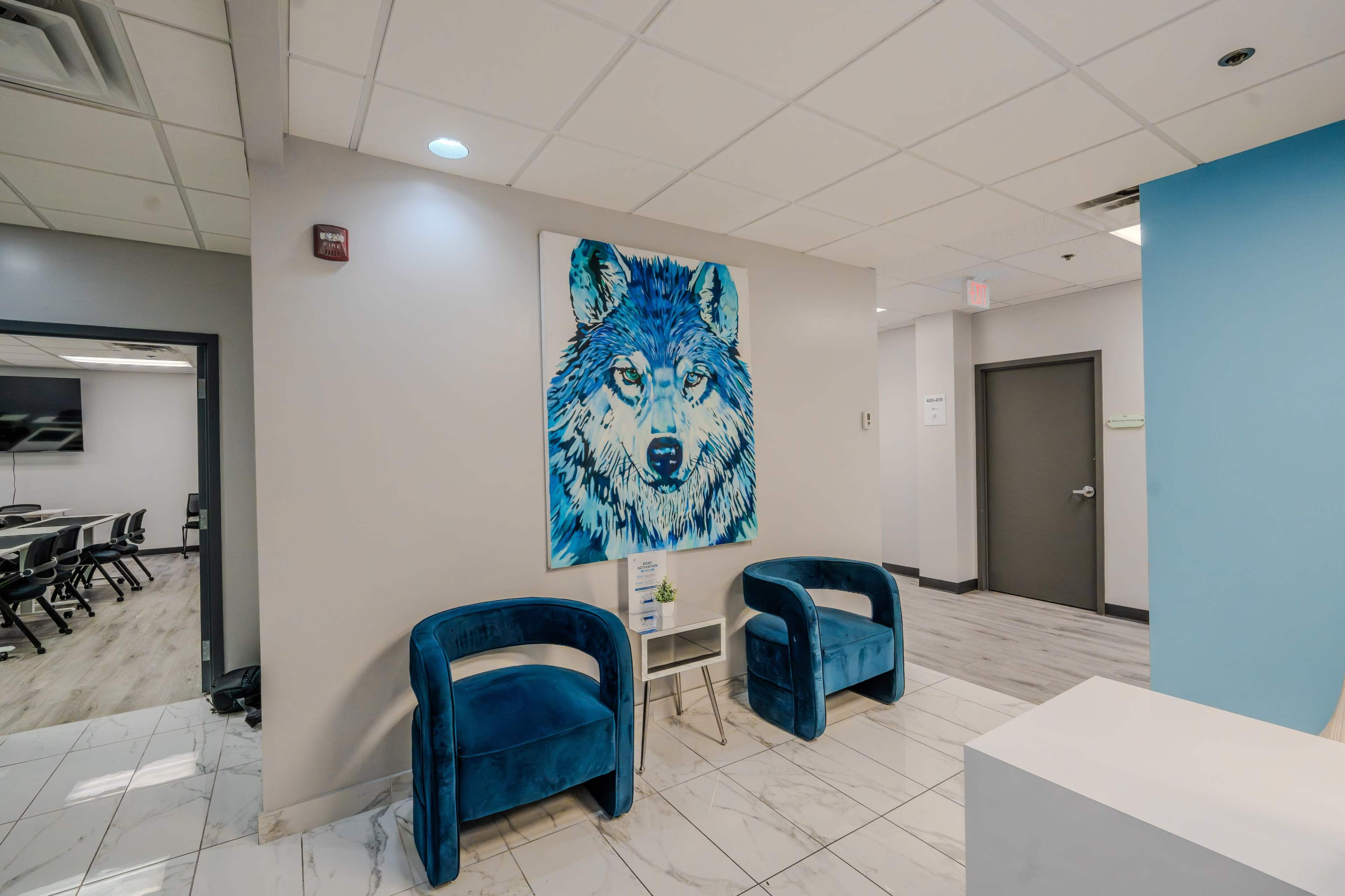 The image shows a modern waiting area featuring two blue chairs, a small table, and a large wolf painting on the wall.