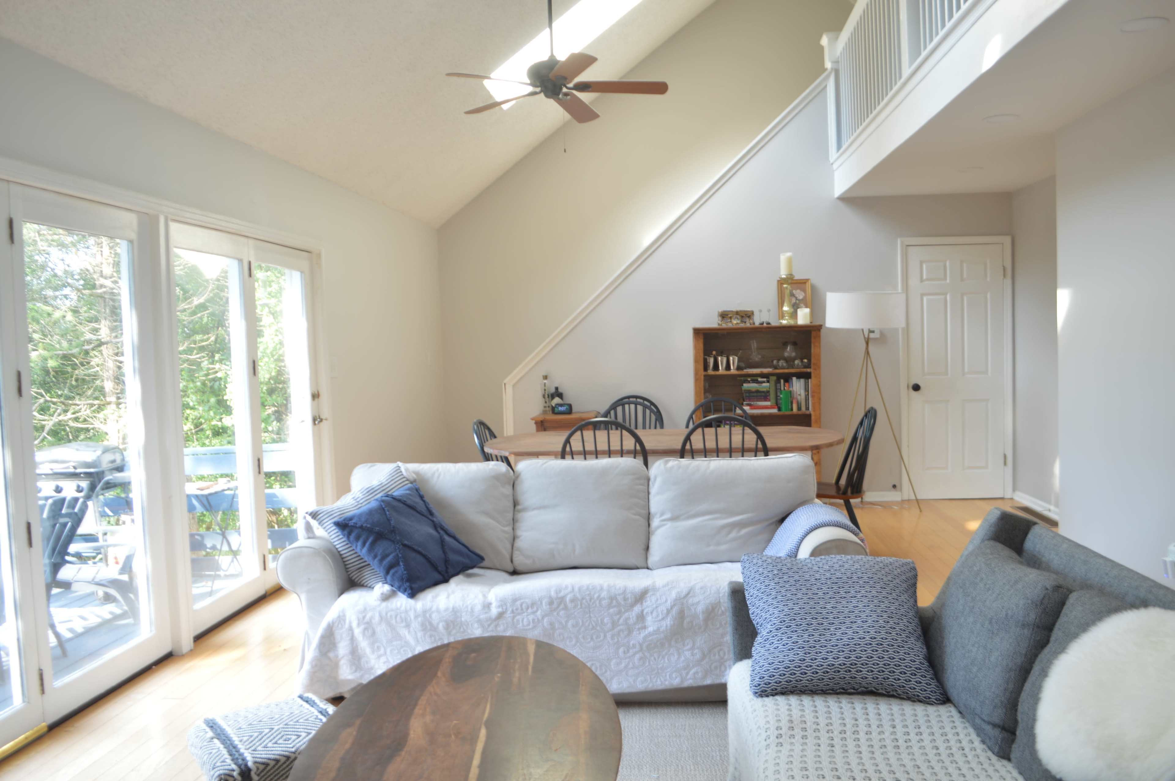 A bright living room features a light-colored sofa, a dark sofa, and a dining area with a wooden table and chairs, all illuminated by natural light from large sliding glass doors.