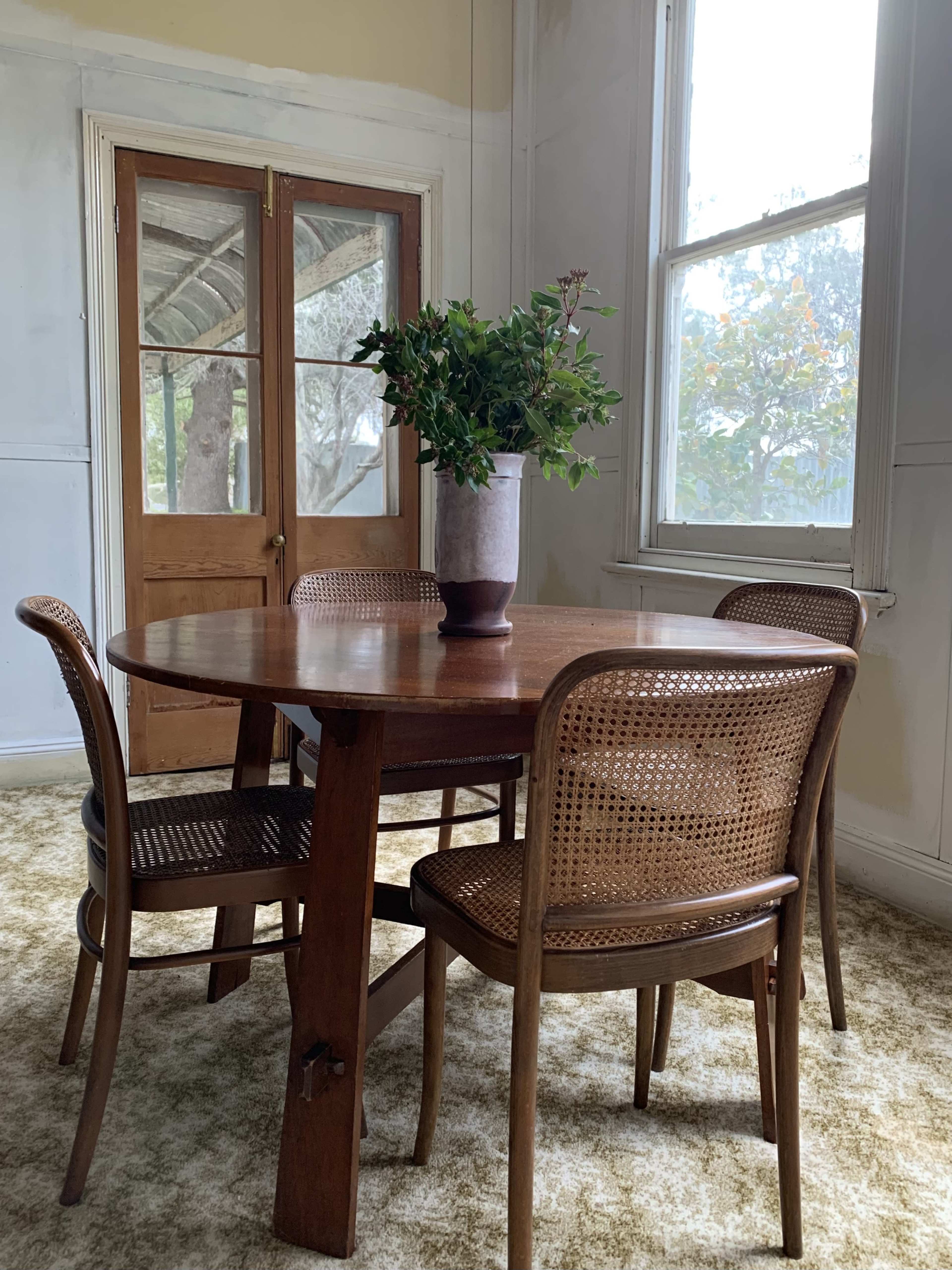 A wooden dining table with four chairs and a vase of greenery sits in a room with large windows and a pair of double doors.