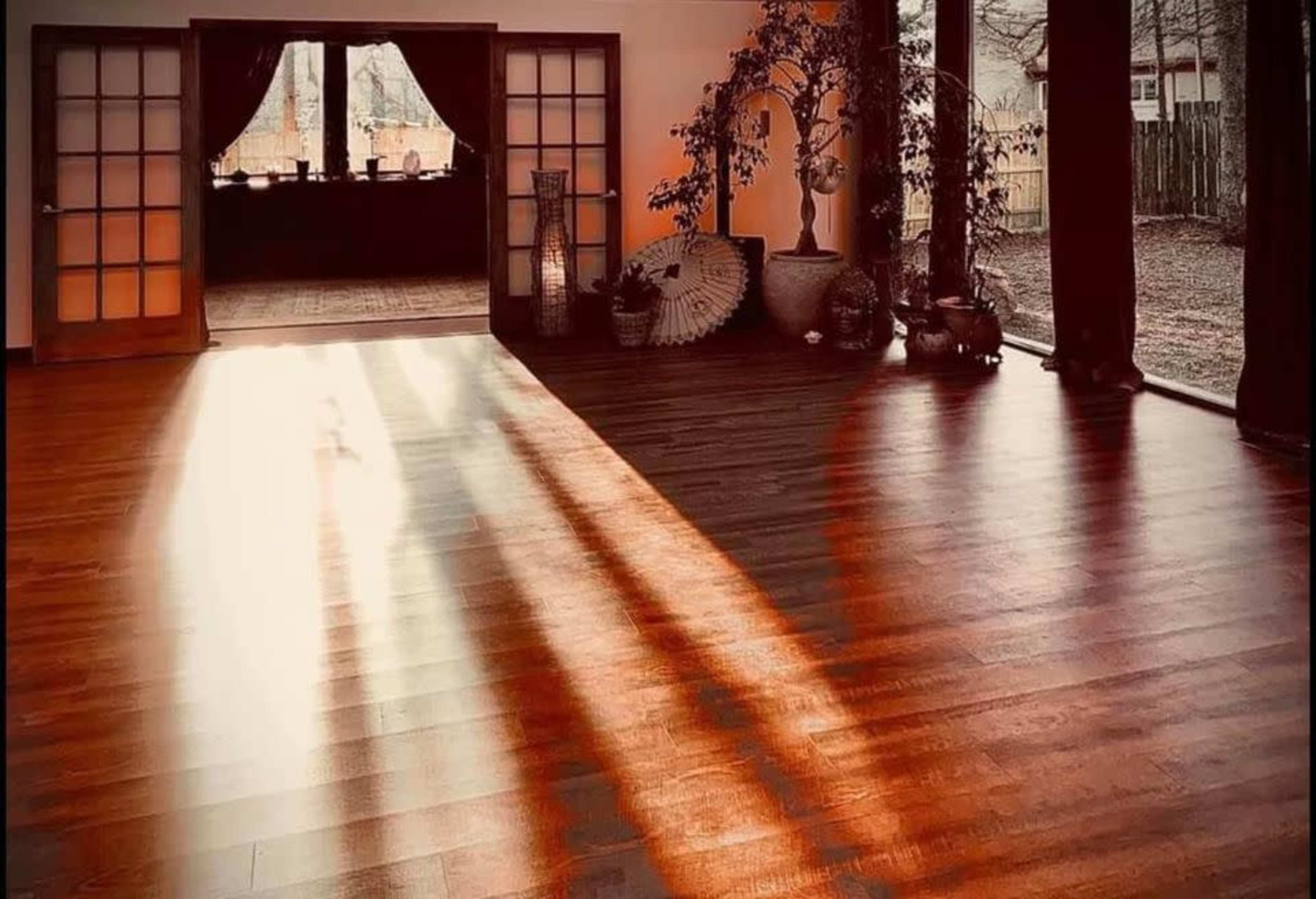 Sunlight streams through large windows onto a wooden floor in a spacious room with plants and decorative elements.