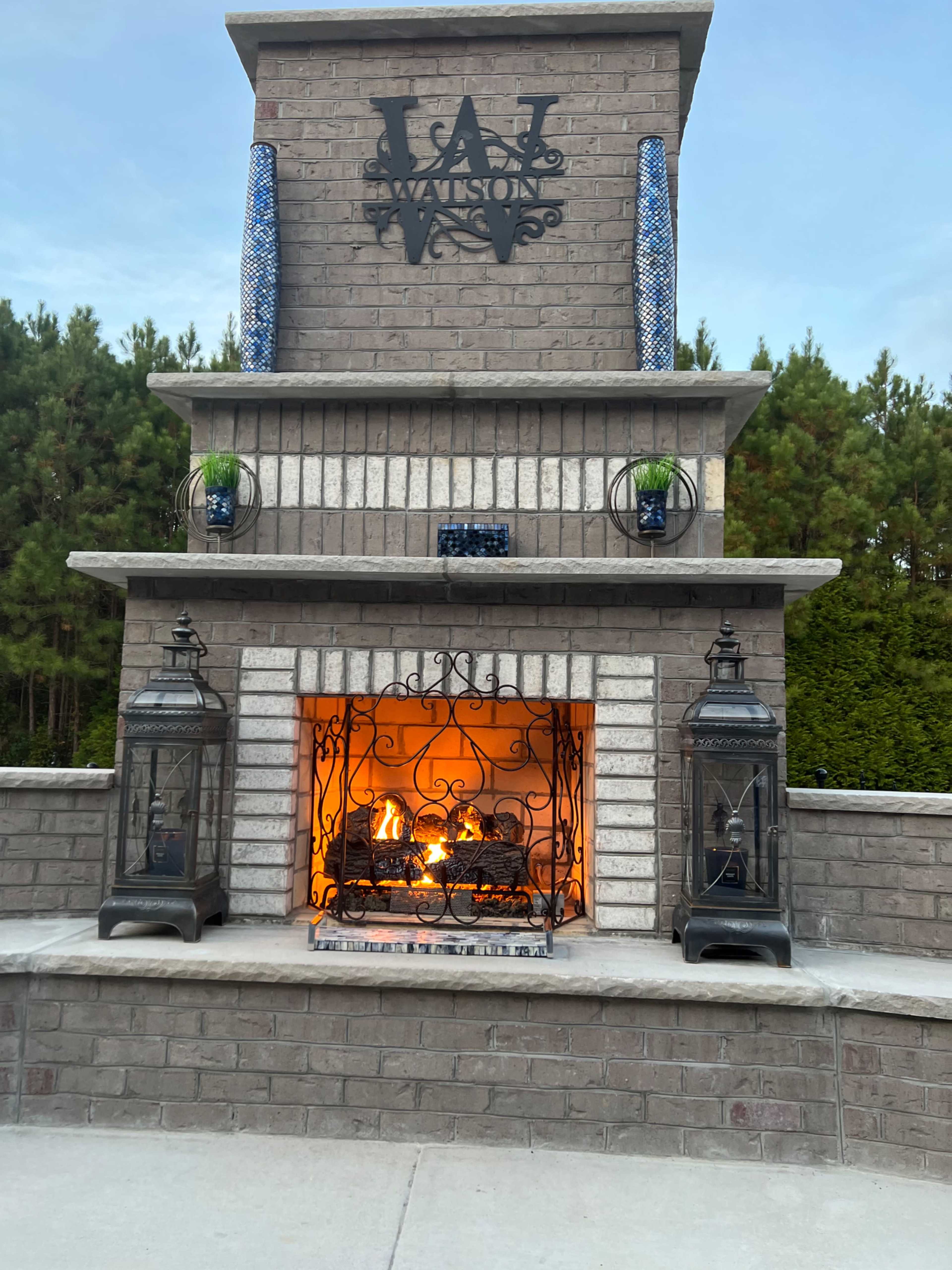 An outdoor stone fireplace with a lit fire, surrounded by decorative lanterns and potted plants.