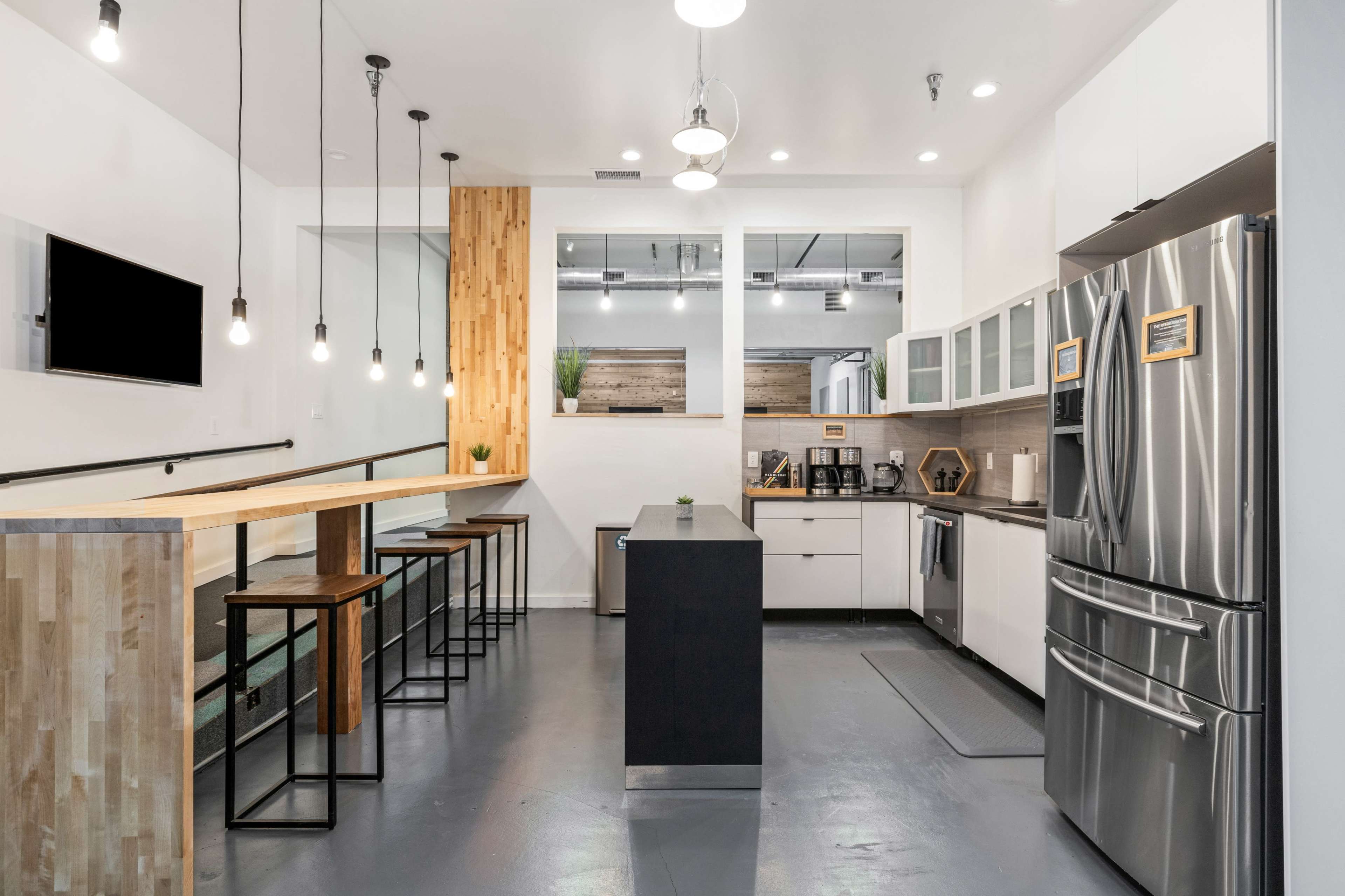 The image shows a modern kitchen featuring stainless steel appliances, wood accents, a long countertop with bar stools, and pendant lighting.