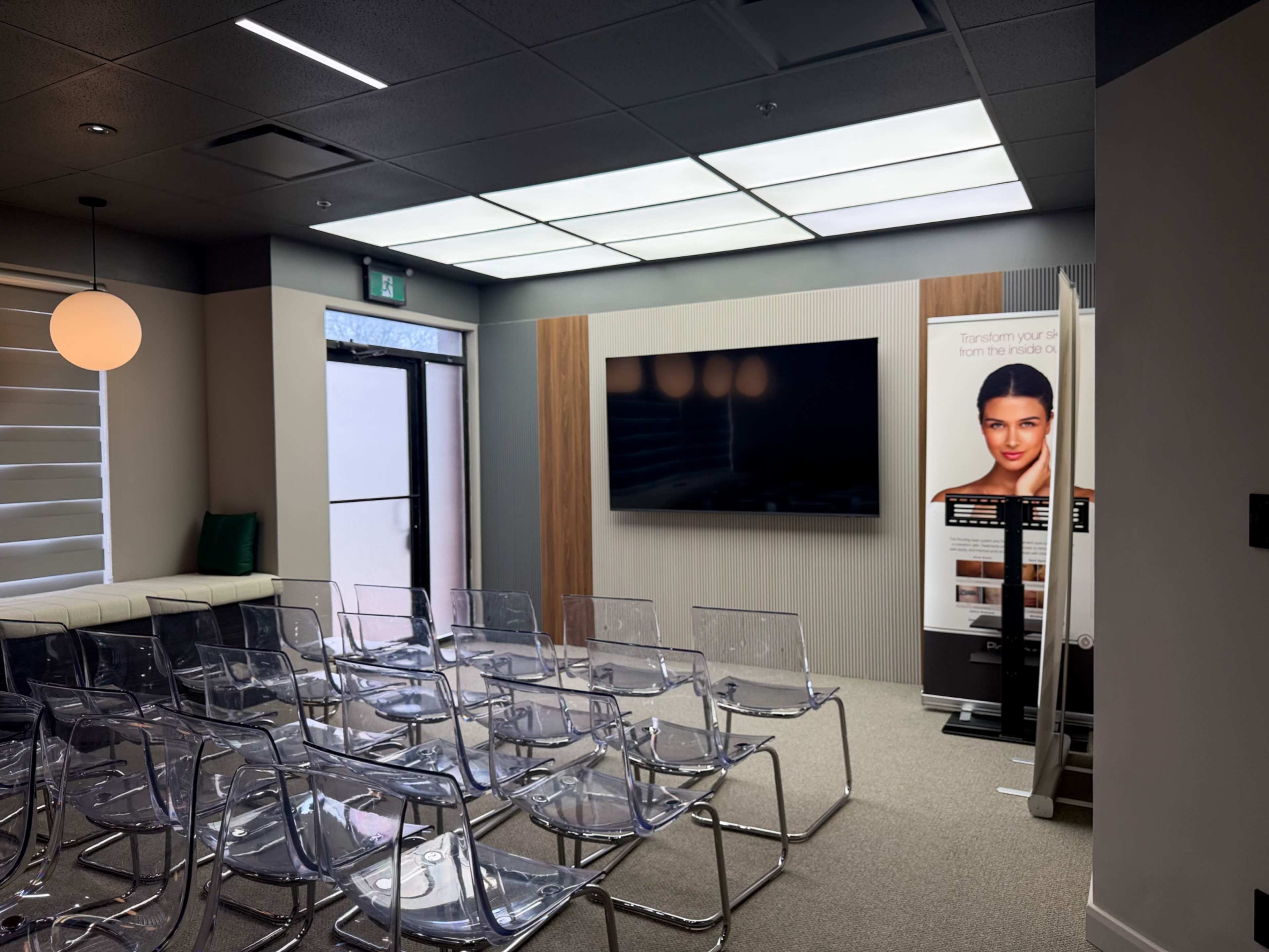 The image shows a modern, minimalist meeting room with rows of transparent chairs, a large screen on the wall, and a poster displaying a woman's face and a transformation message.