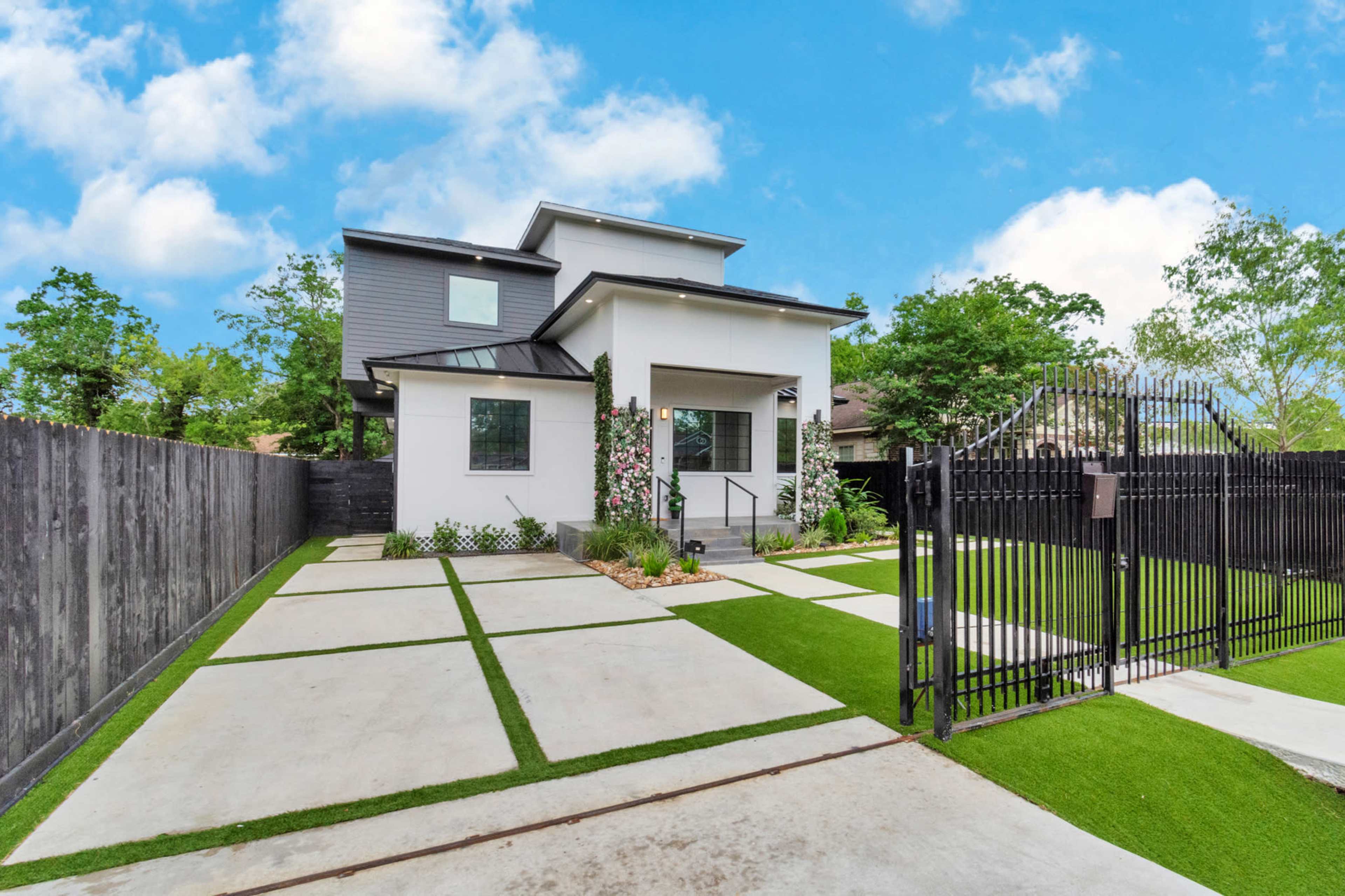 A modern house with a clean design is set behind a black gated entrance, featuring a neatly landscaped front yard with concrete paths and artificial grass.