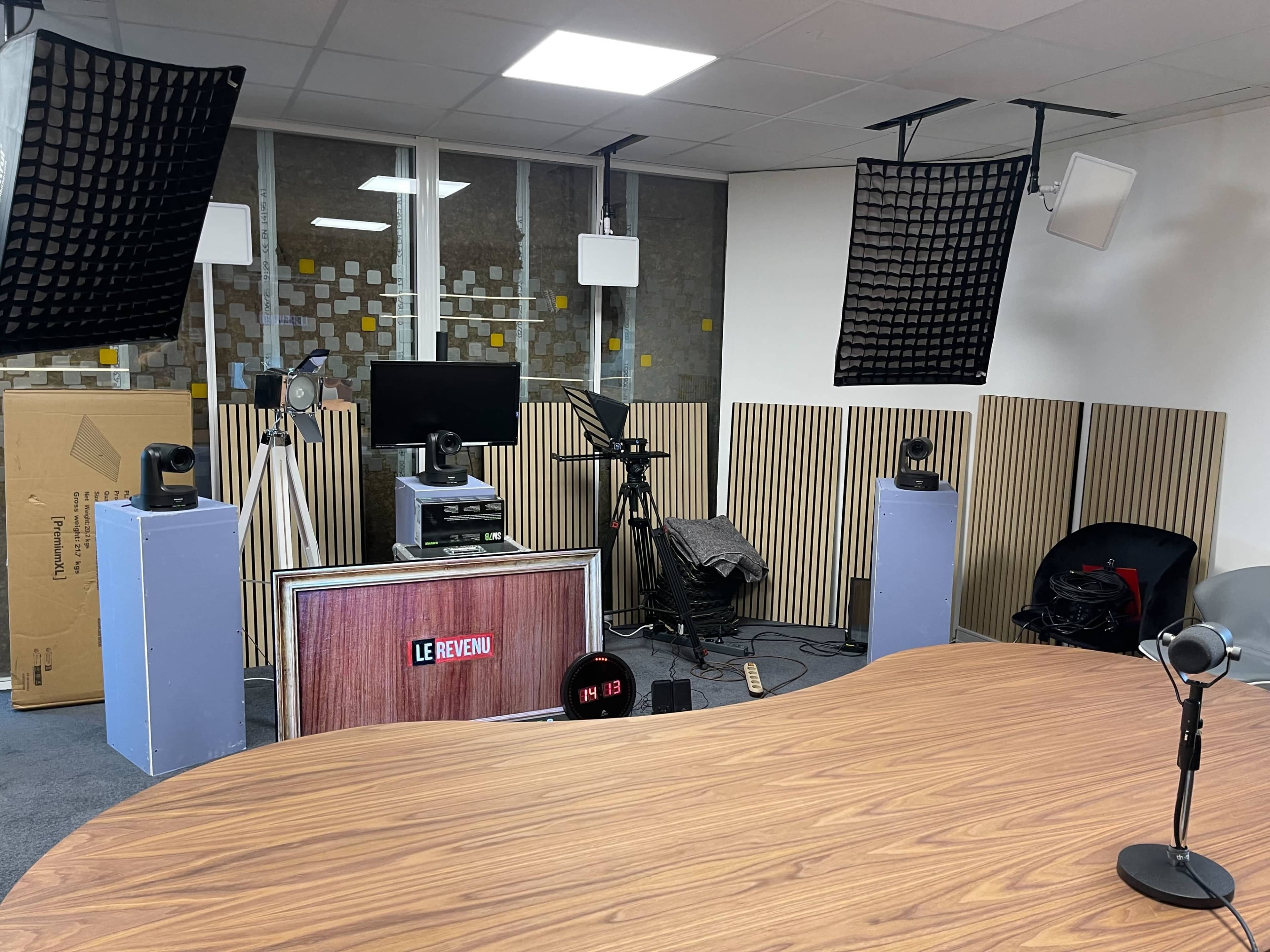 The image shows a modern recording studio with a large wooden table, multiple cameras, microphones, and sound equipment arranged in a well-lit space.