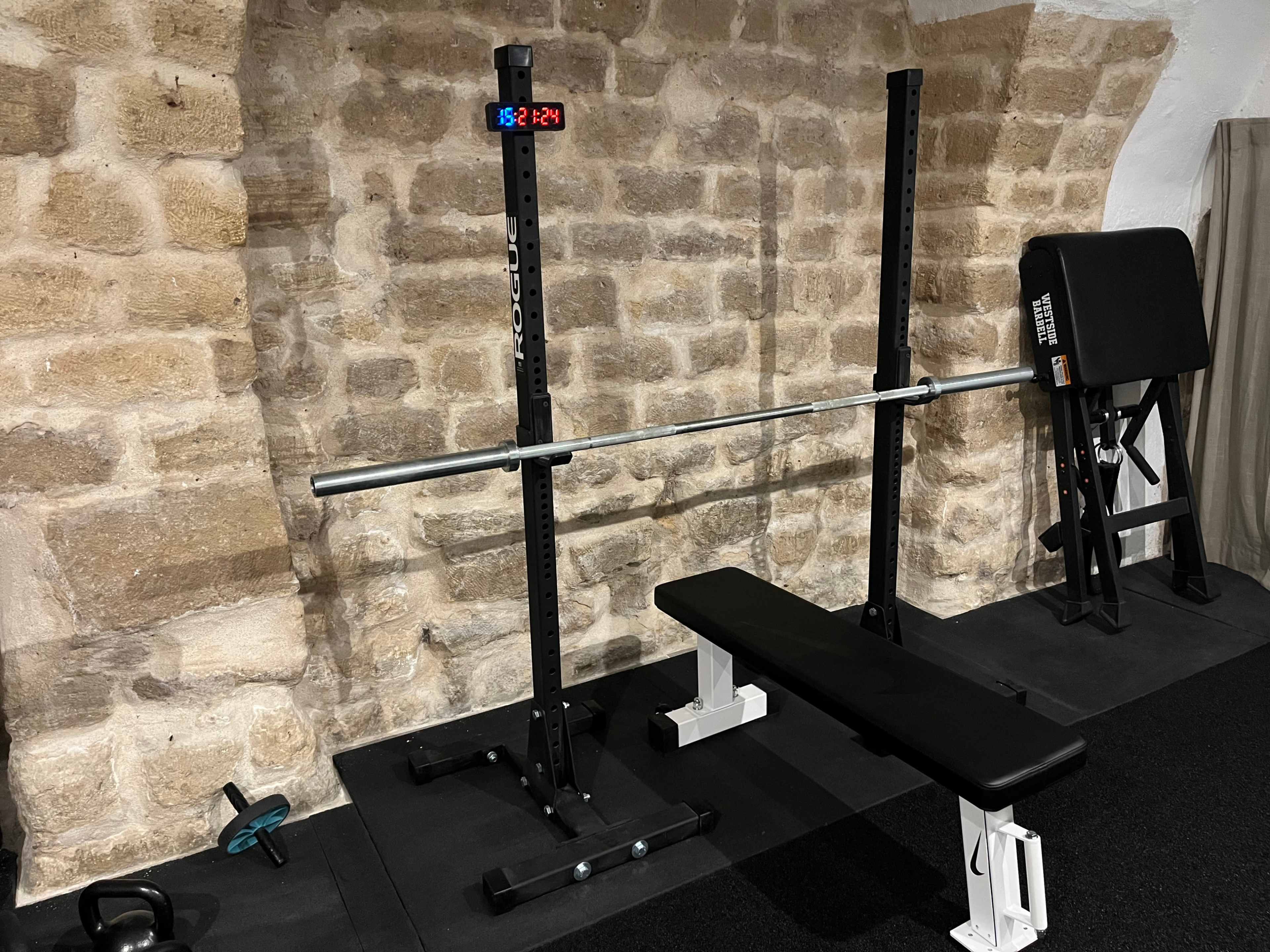 The image shows a gym setup featuring a weight bench, a barbell on a squat rack, and a digital timer mounted on the wall.