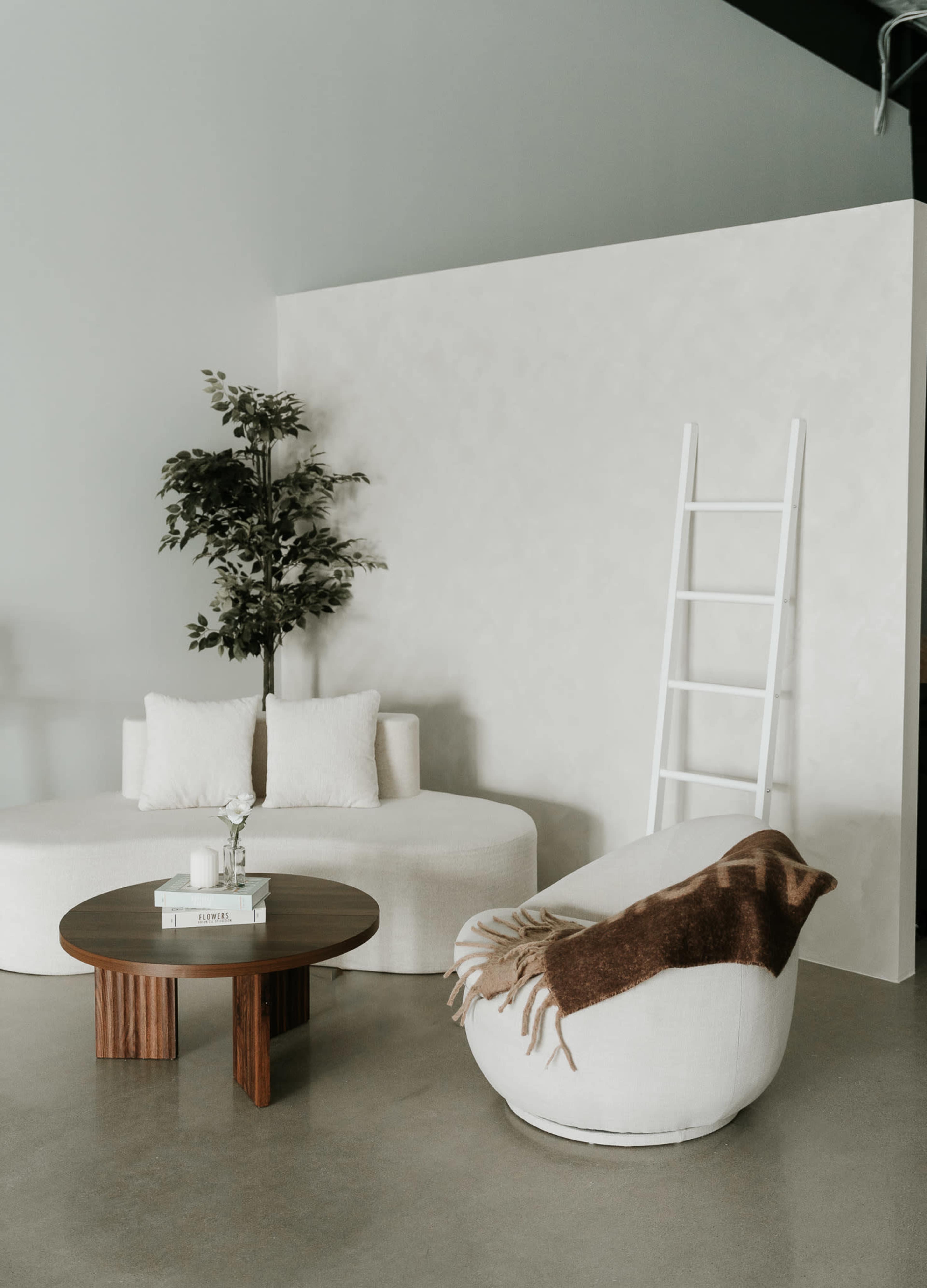 The image shows a modern living space featuring a round white sofa, a wooden coffee table, a cozy floor pouf, and a decorative ladder leaning against a textured wall.