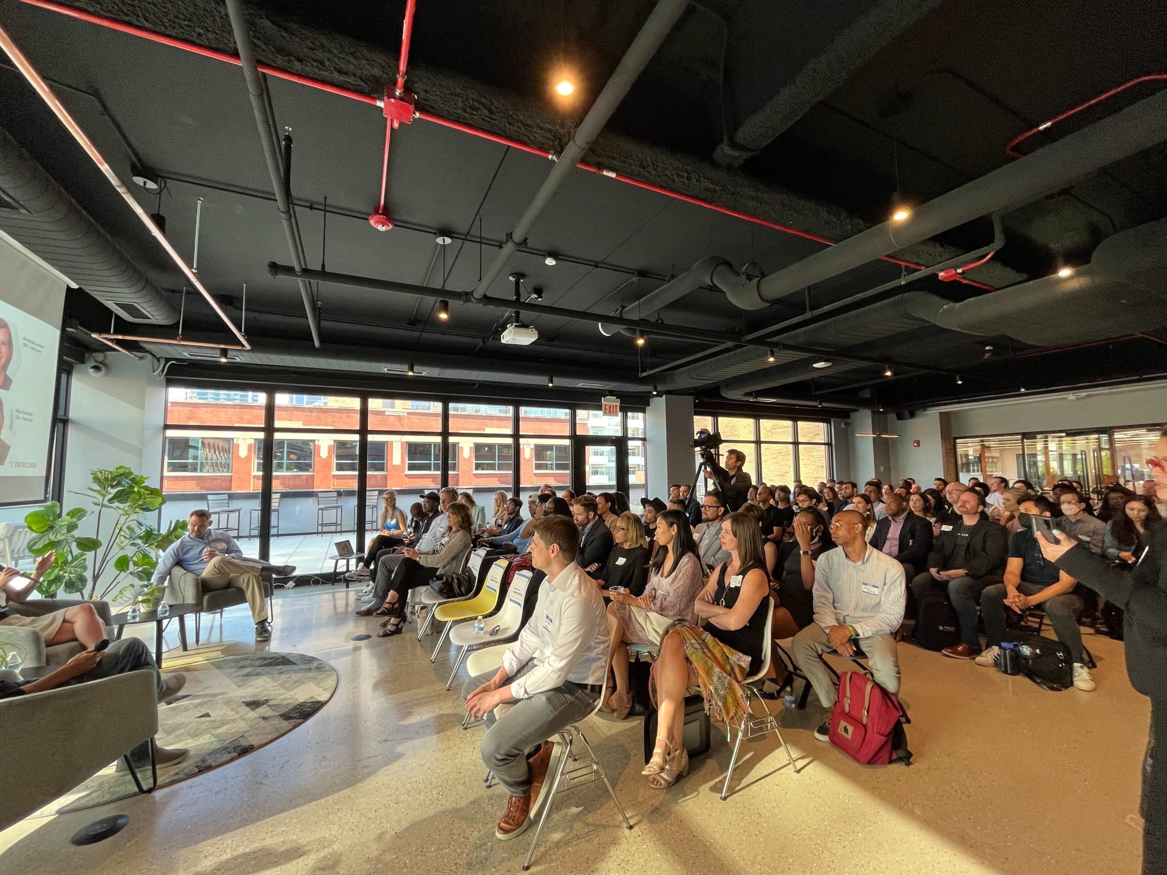 A large audience is seated in a modern conference room, attentively watching a presentation on a screen.