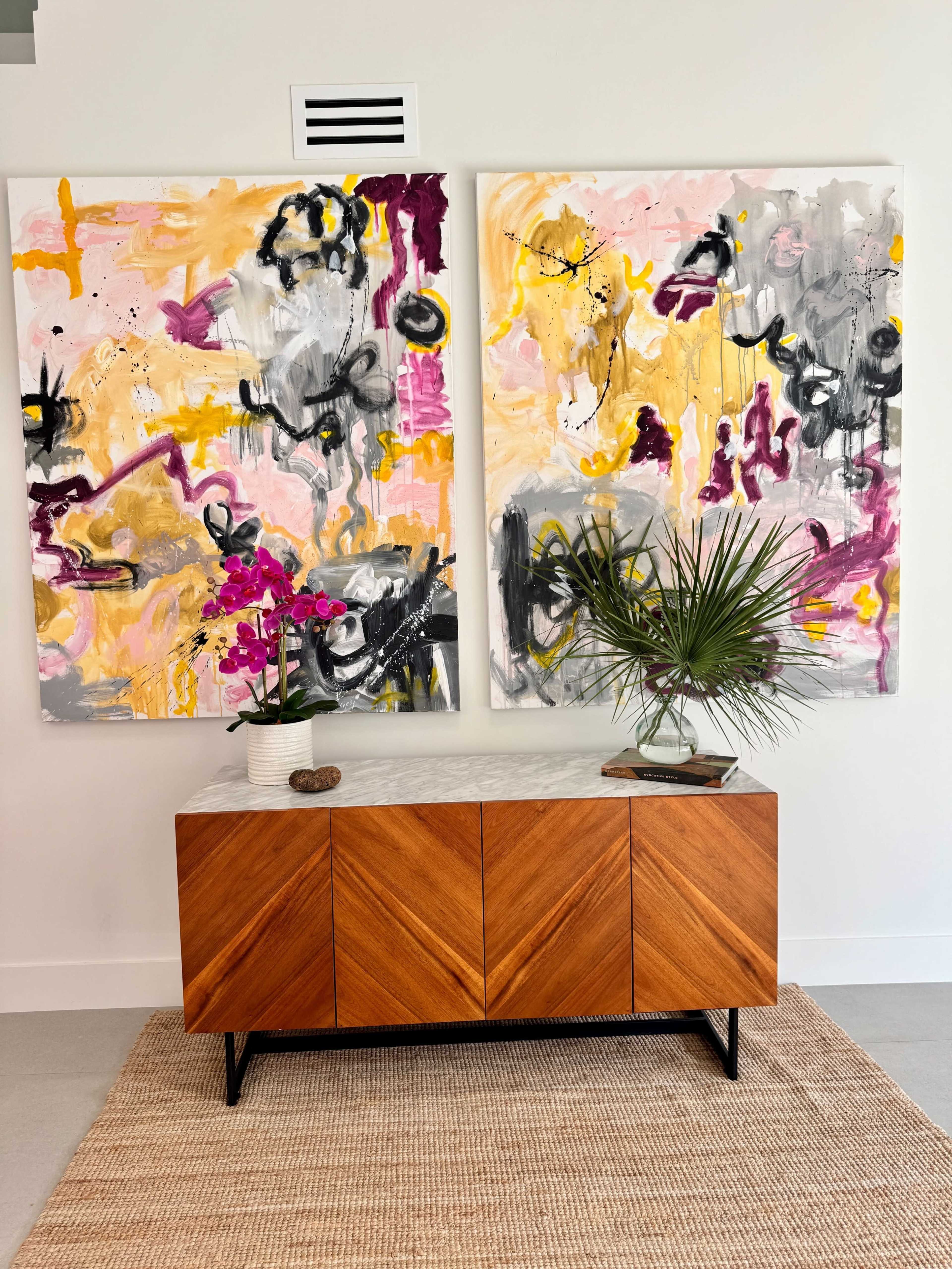 The image features a wooden sideboard with a marble top, placed on a woven rug, accompanied by two large abstract paintings in vibrant colors and a decorative arrangement of plants and flowers on top.