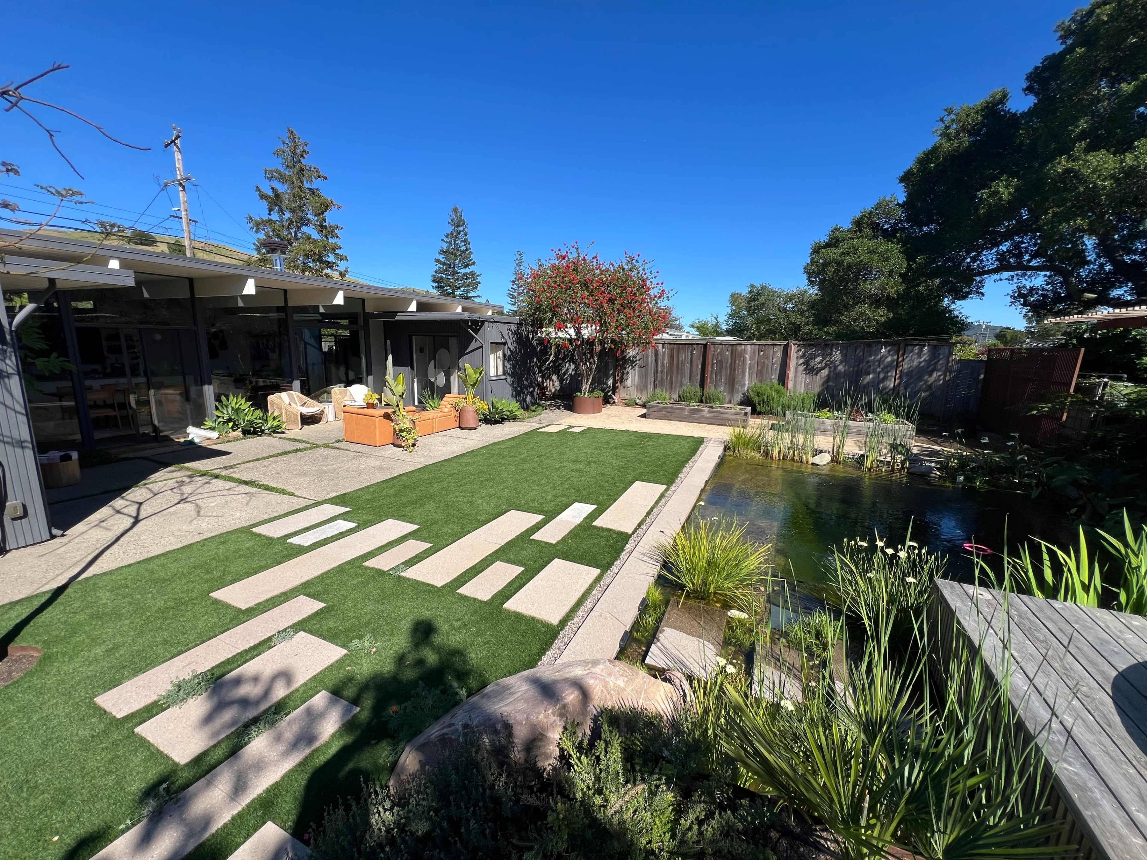 The image shows a landscaped garden with a pond, stone pathways, and a modern home in the background.