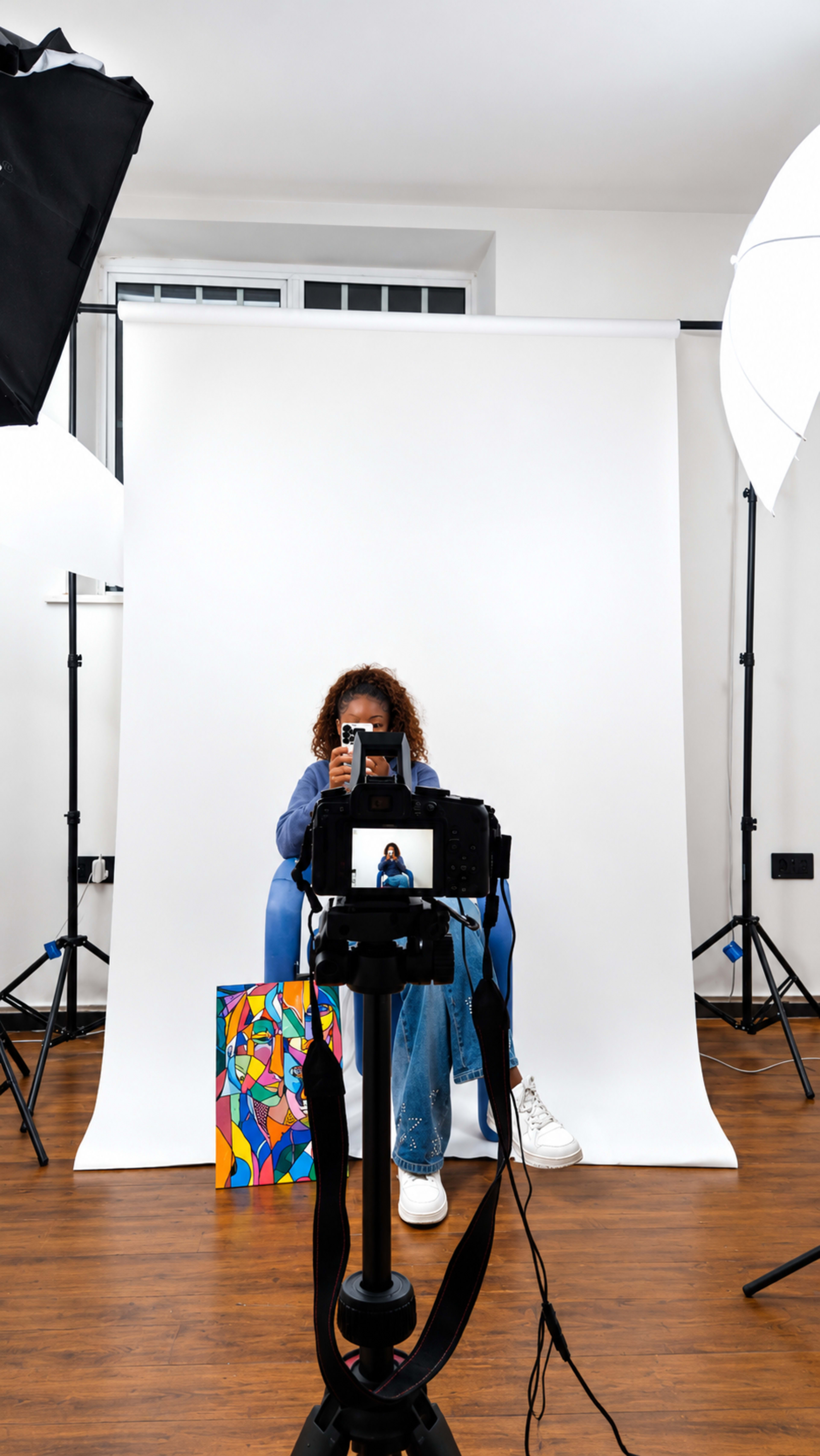 A person with curly hair is seated in front of a white backdrop, holding a camera while a colorful bag rests beside them, surrounded by studio lighting setup.