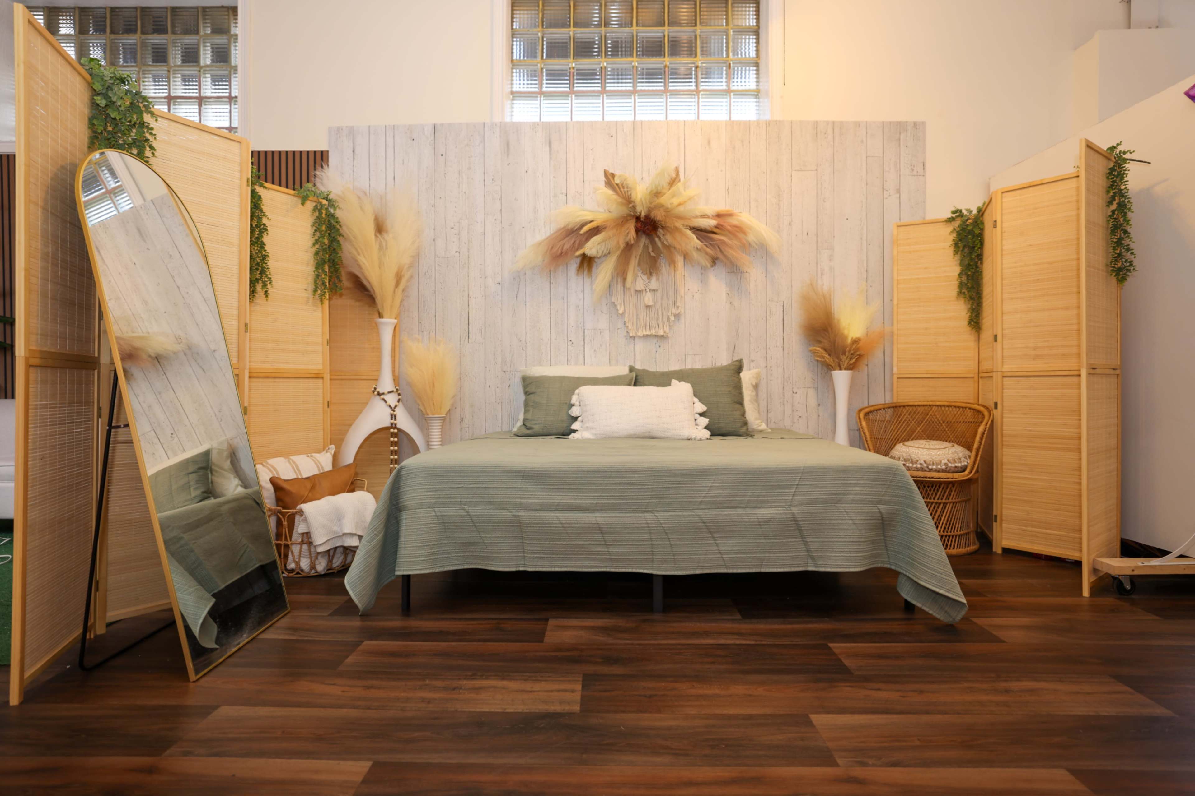 The image depicts a bedroom with a bed covered in a green blanket, flanked by decorative screens, a large mirror, and various plant accents.