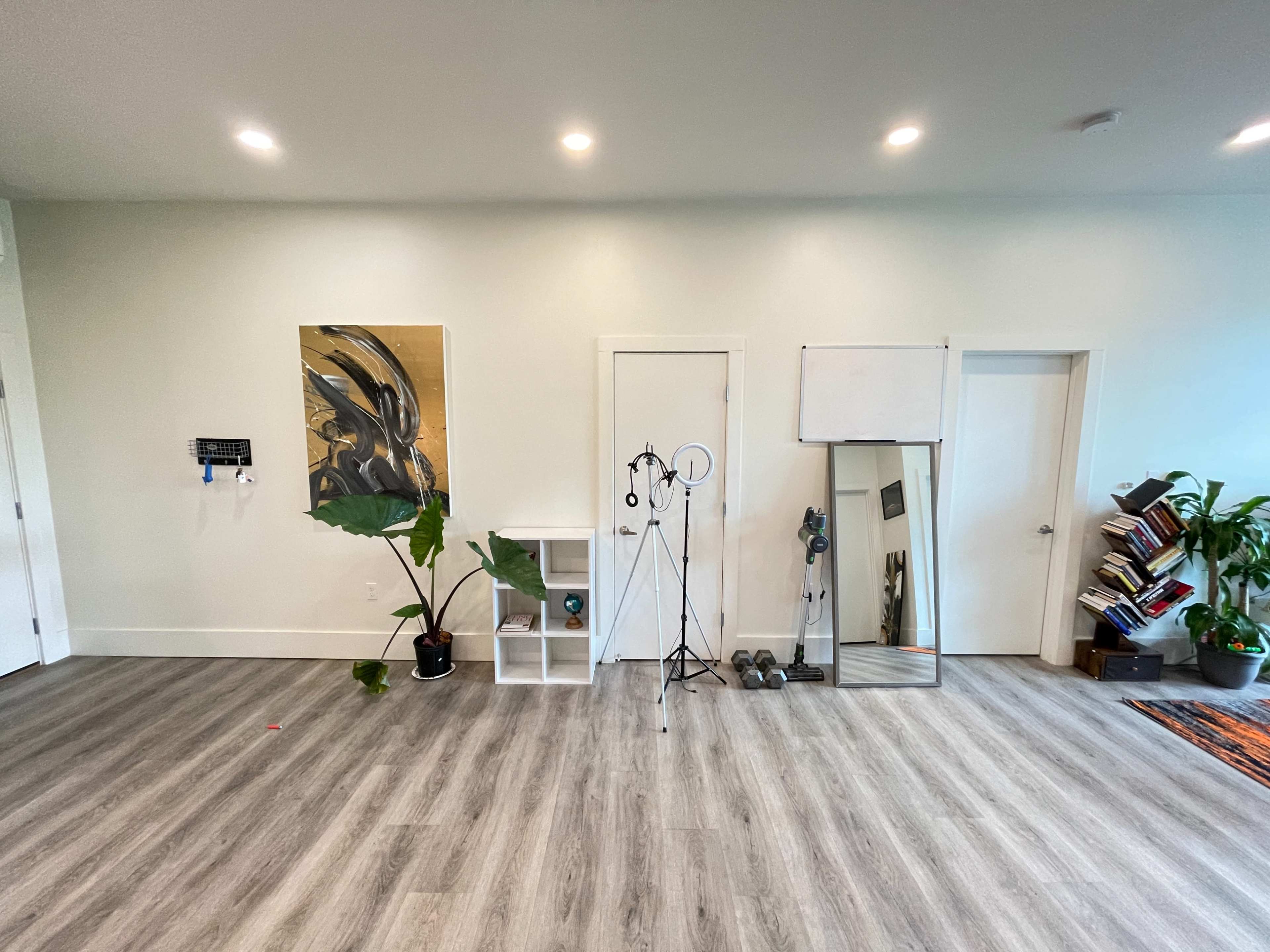 A spacious room with light-colored walls, wooden flooring, a plant, a shelf, a mirror, and various fitness equipment.