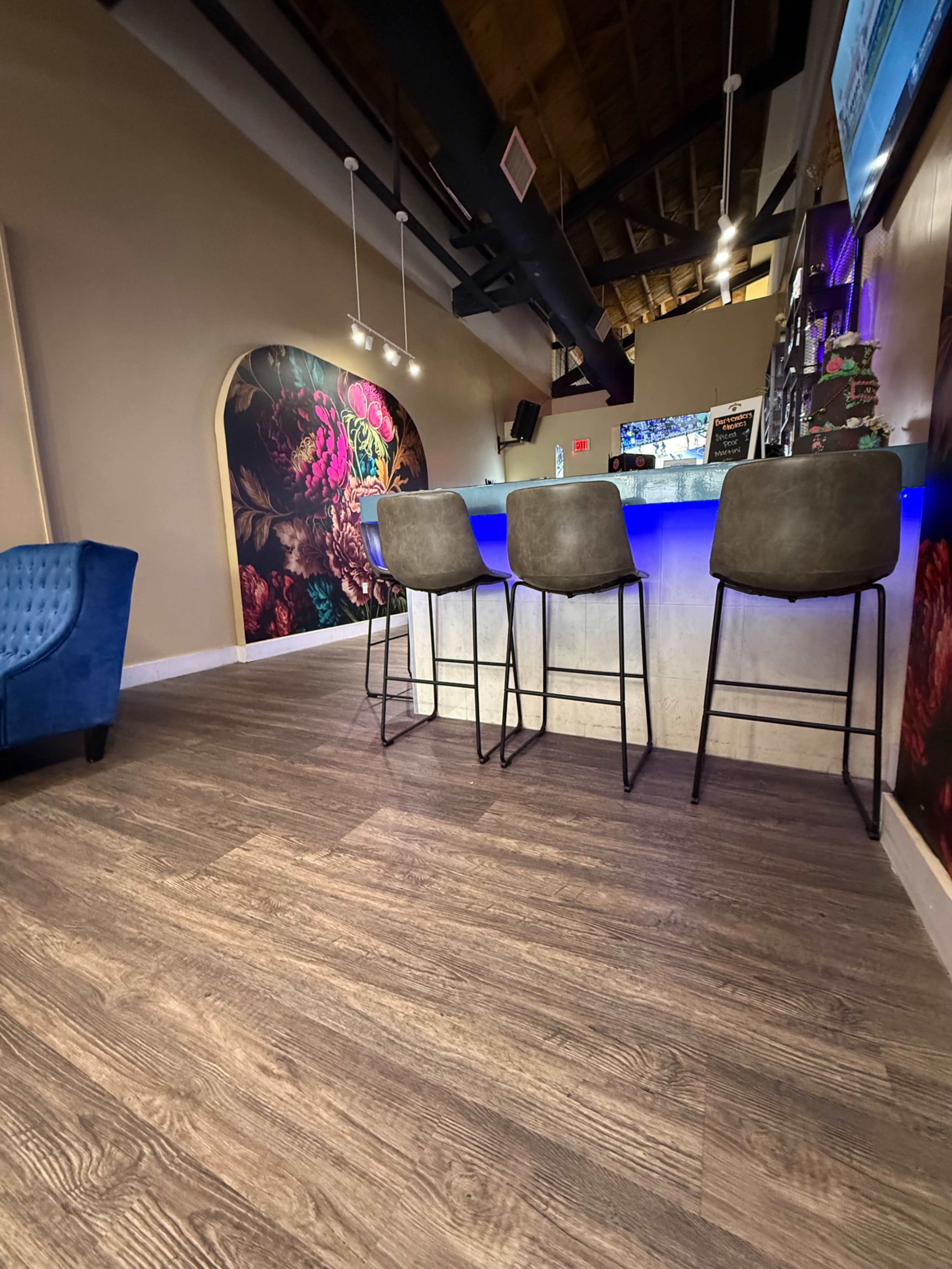 The image shows a modern, inviting bar area with four high stools positioned in front of a brightly lit counter and colorful wall art.