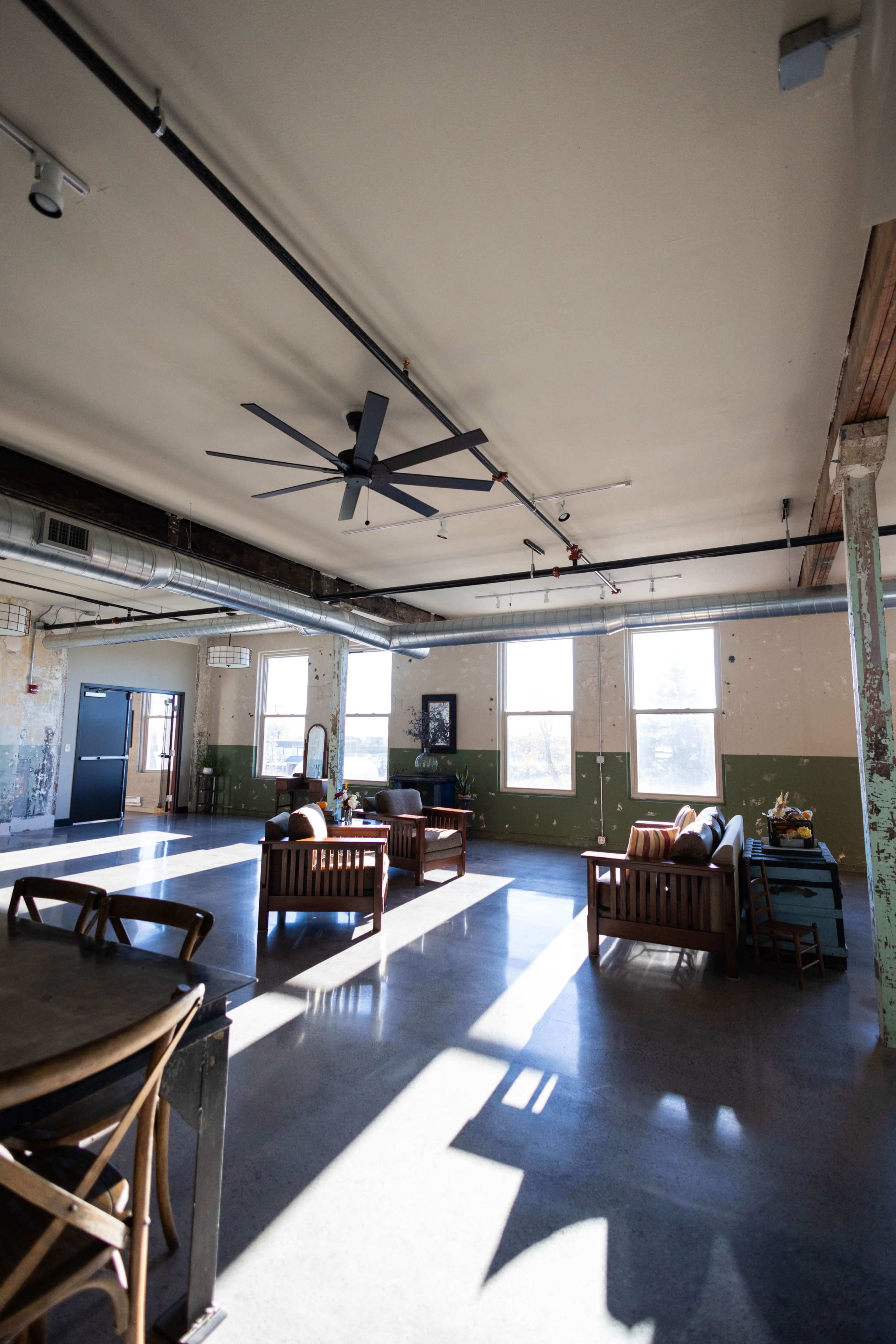 A spacious, industrial-style room with wooden seating, large windows, and a ceiling fan.