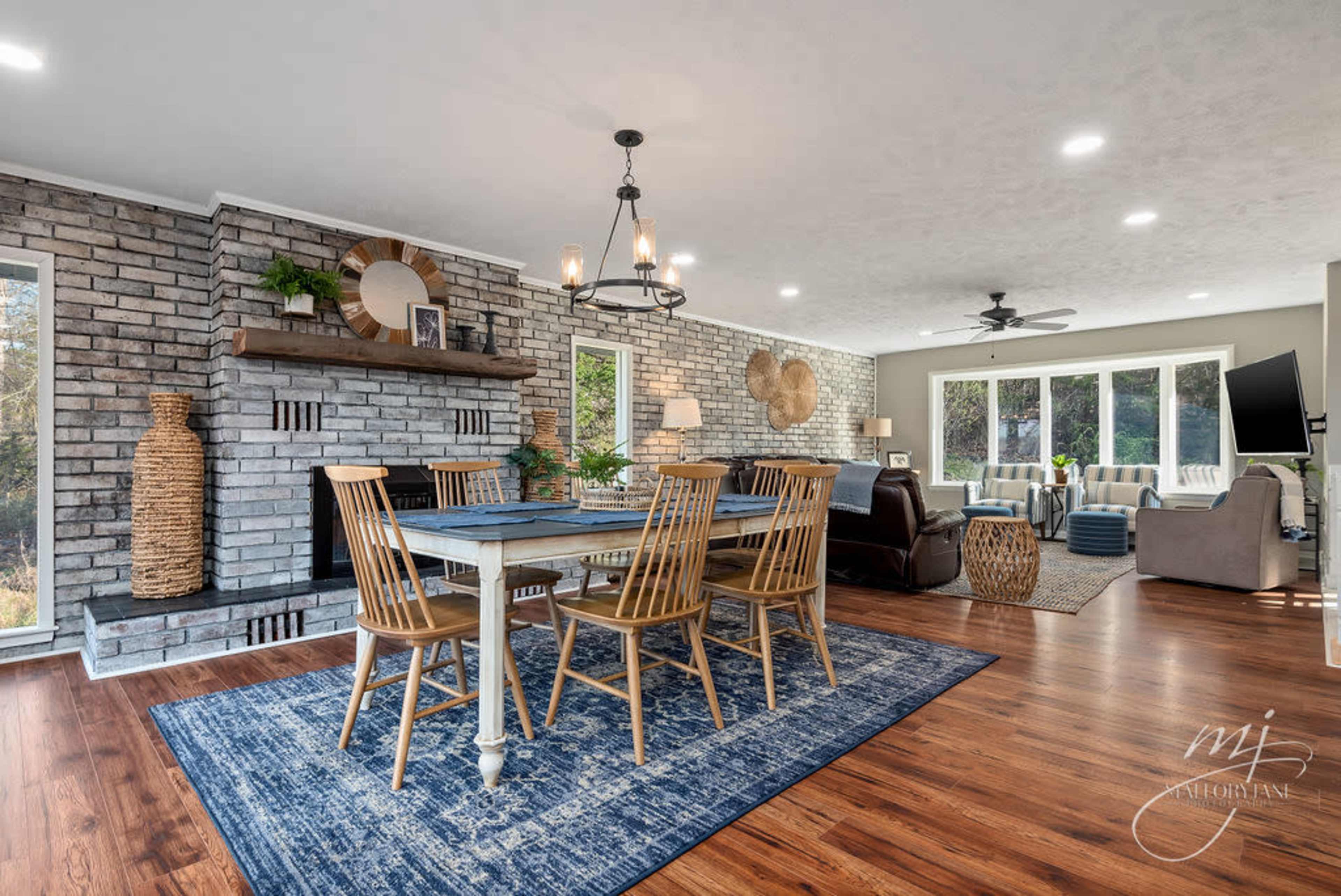 The image shows a modern living and dining area featuring a wooden dining table, a stone fireplace, and large windows, with a combination of stylish decor elements and comfortable seating.