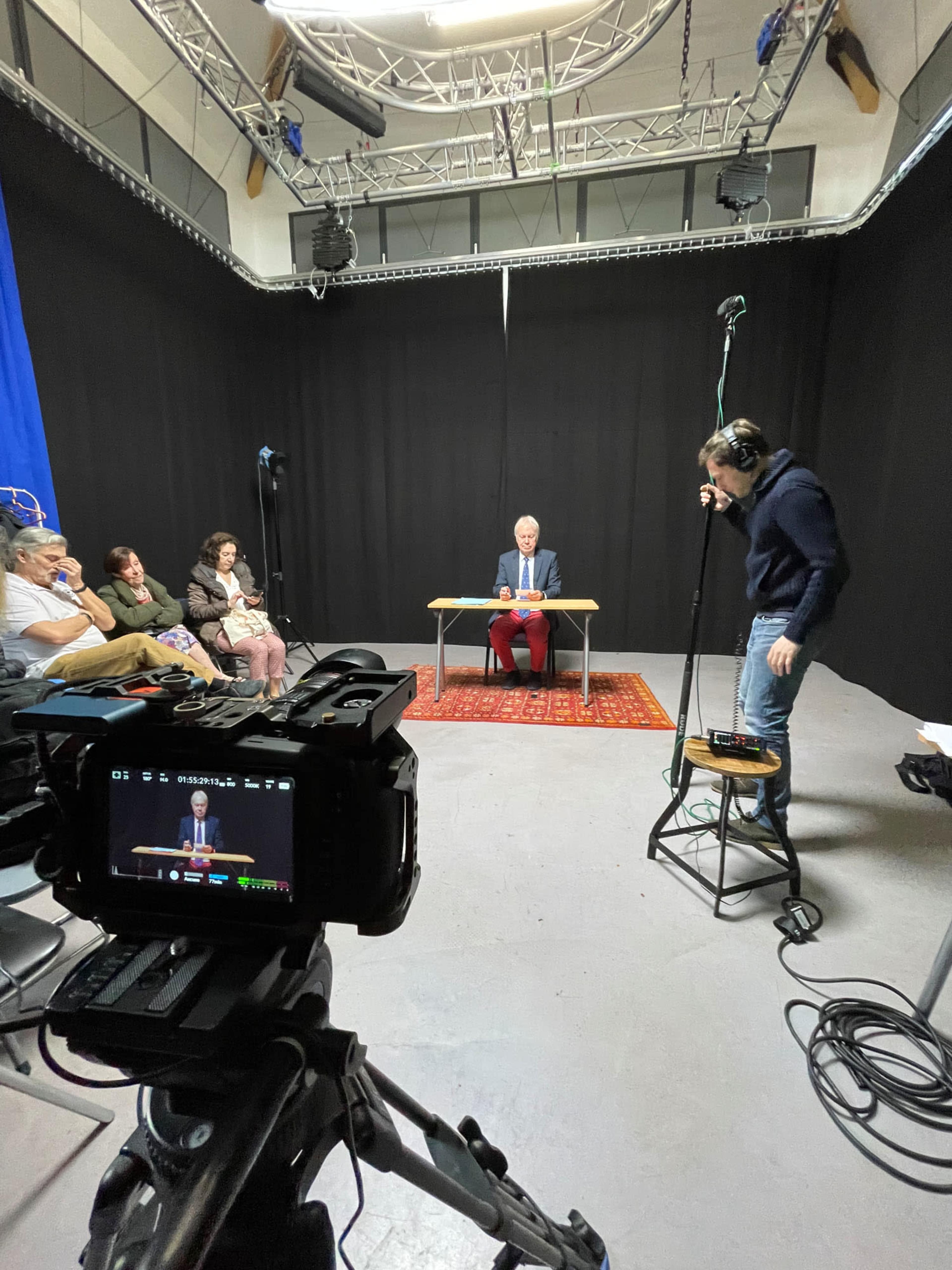 A man in a suit sits at a table while a technician adjusts equipment in a dimly lit studio, and an audience observes from the side.