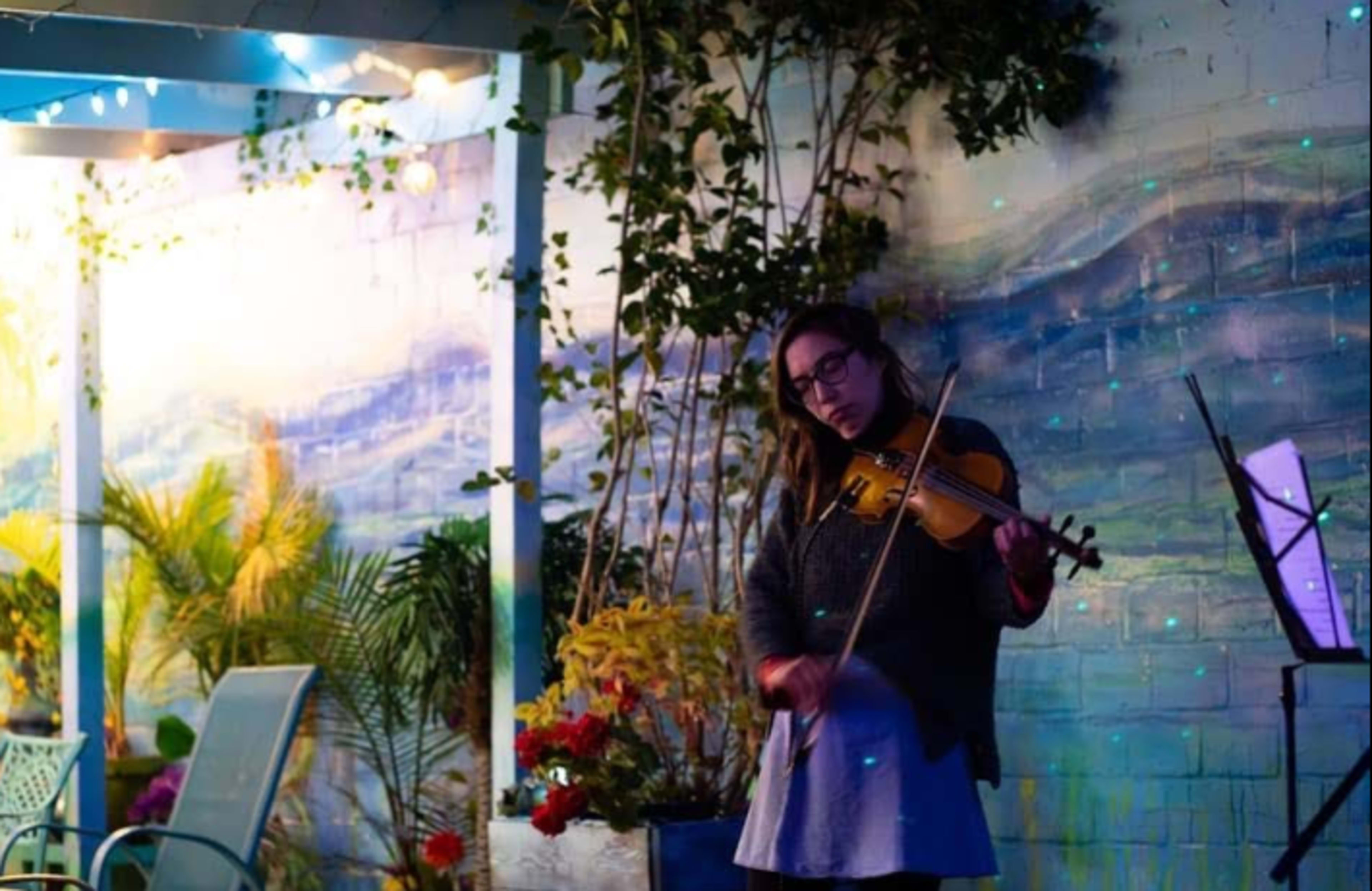 A woman plays a violin in front of a painted mural surrounded by plants and colorful lighting.