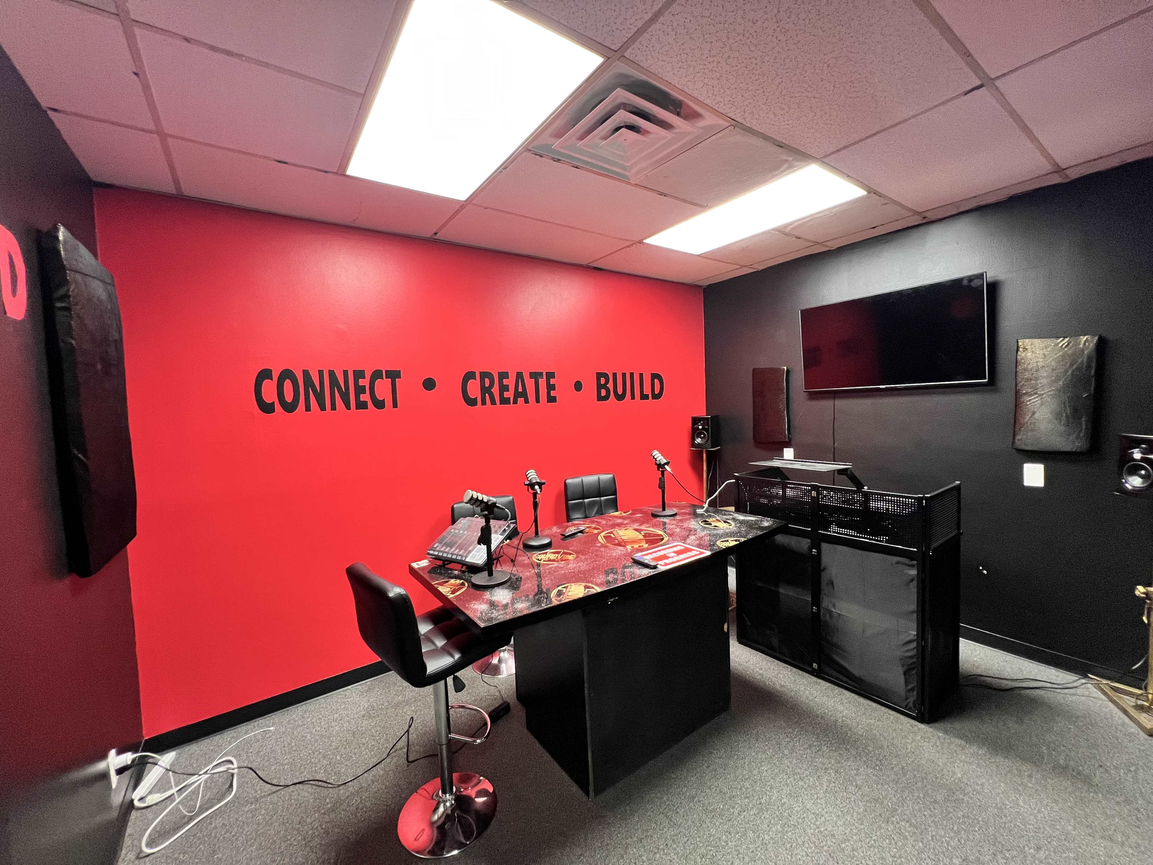 A recording studio with a red and black color scheme, featuring microphones on a table, chairs, and a television mounted on the wall.