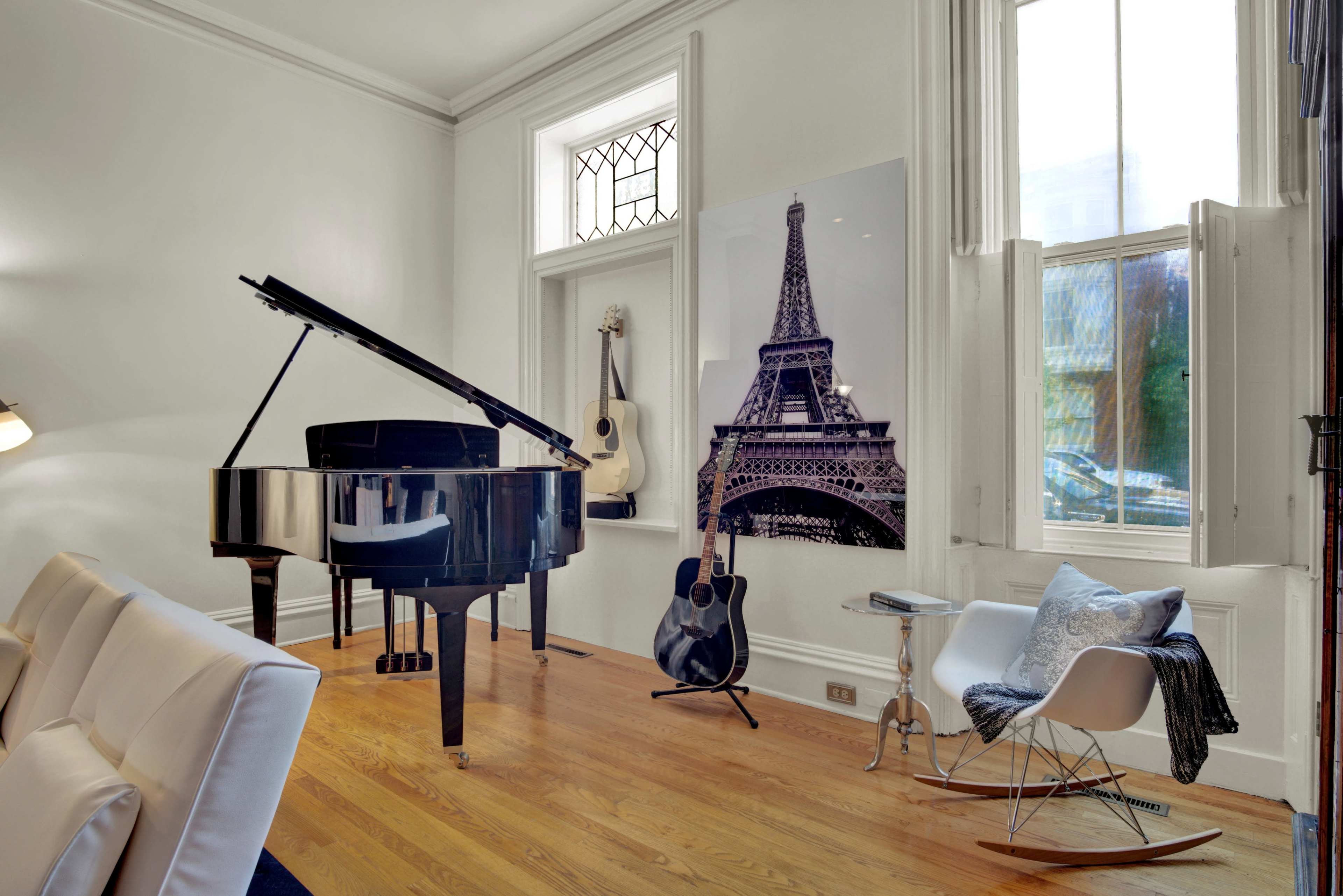 A black grand piano sits next to an acoustic guitar and a photograph of the Eiffel Tower in a sunlit room with large windows.