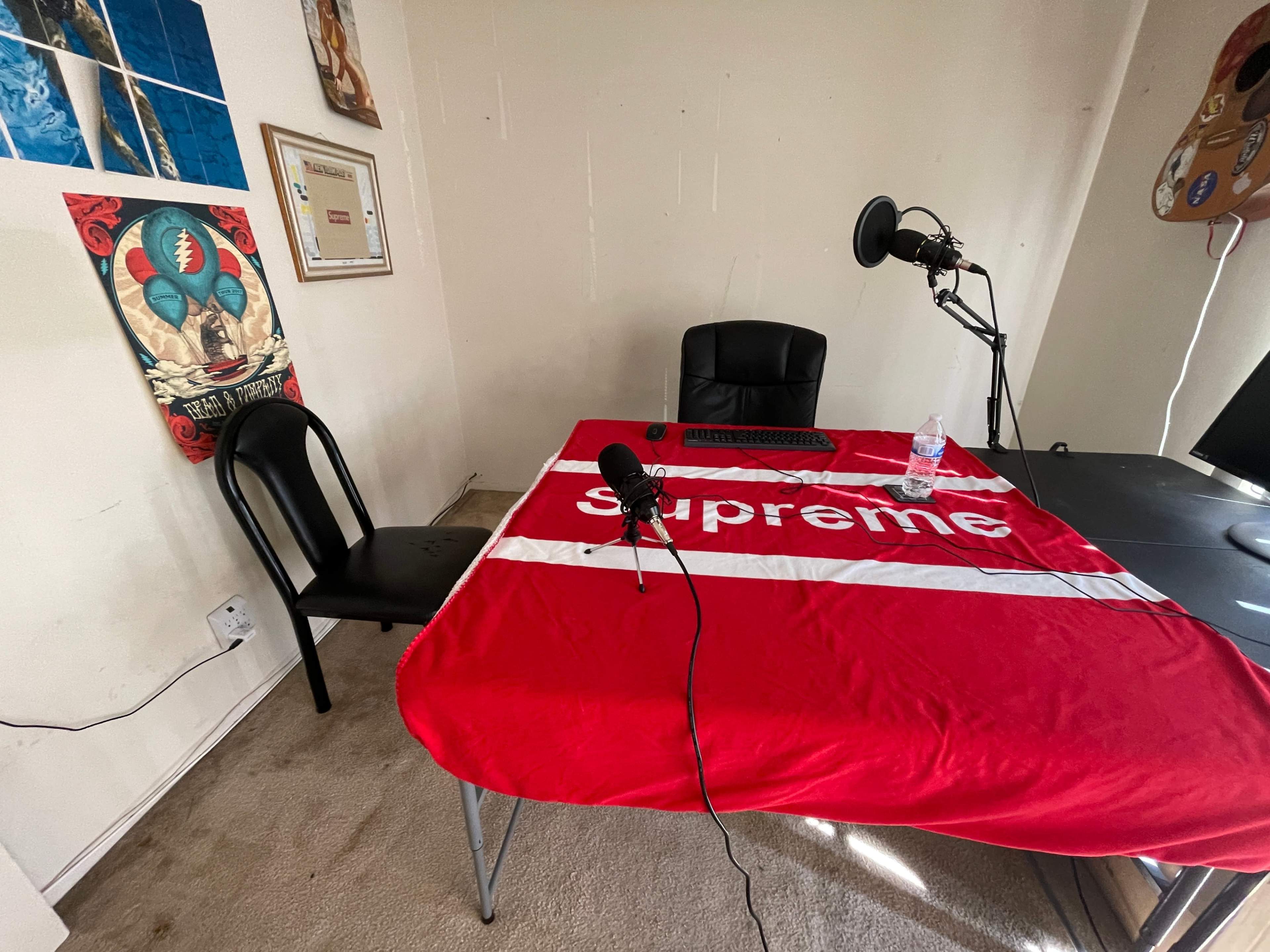 A red tablecloth with "supreme" printed on it covers a table in a room with a black chair, a microphone, and a computer setup.