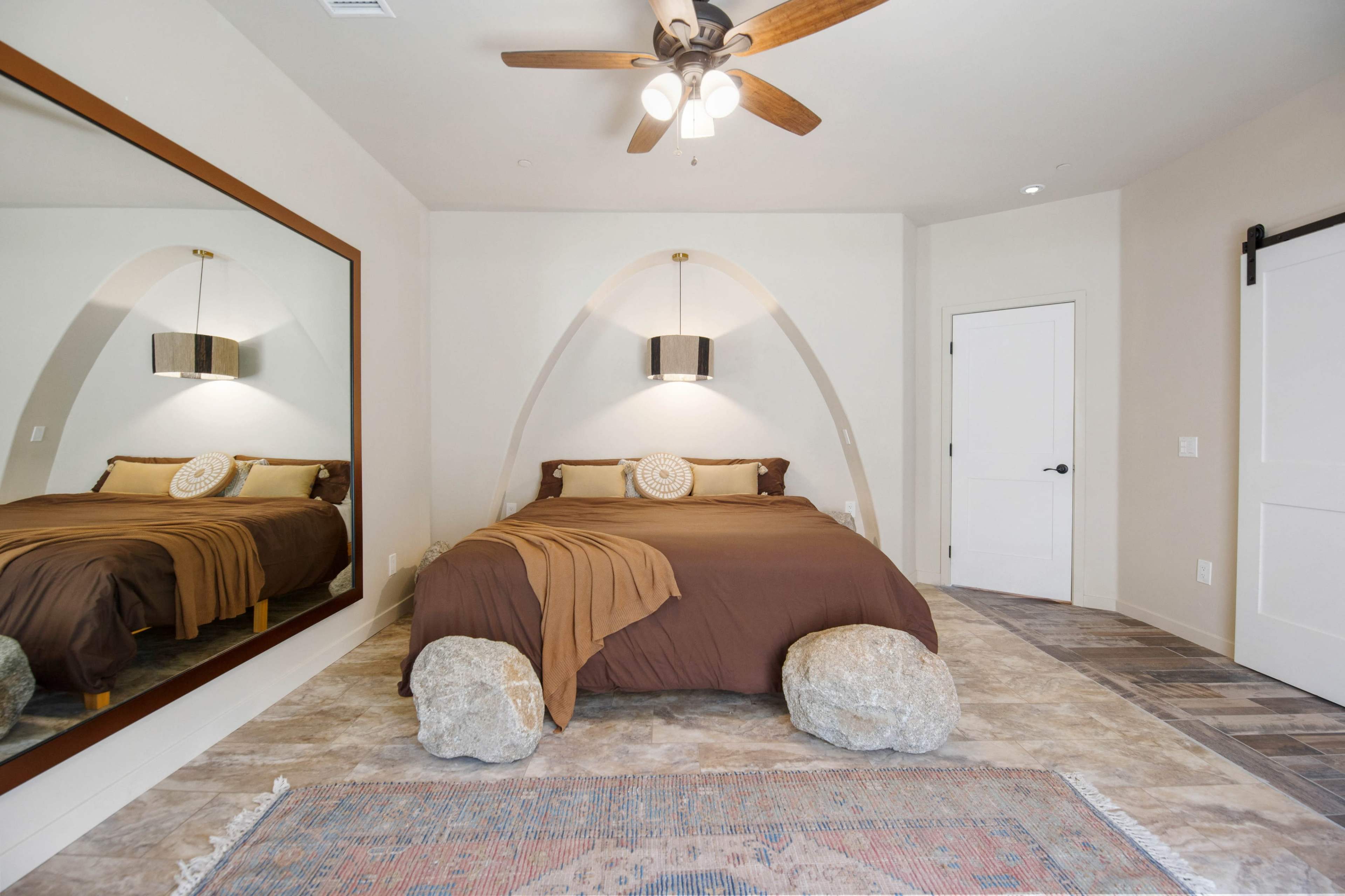 A spacious bedroom features a large bed with brown bedding, a decorative mirror, and two rocks placed at the foot of the bed.
