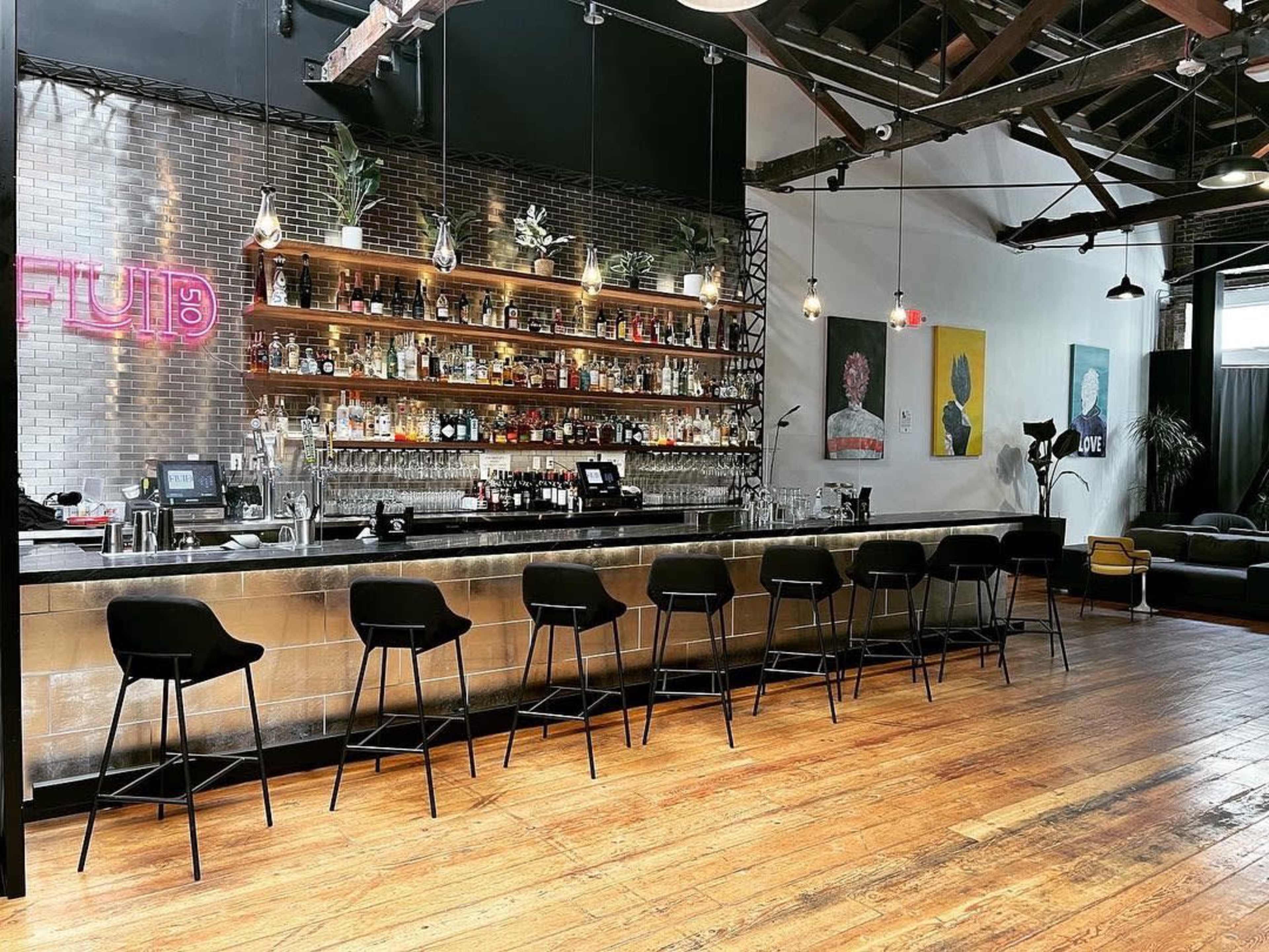 The image shows a modern bar with a wooden floor, a long metallic bar top, and shelves lined with various bottles of liquor, accompanied by black bar stools.