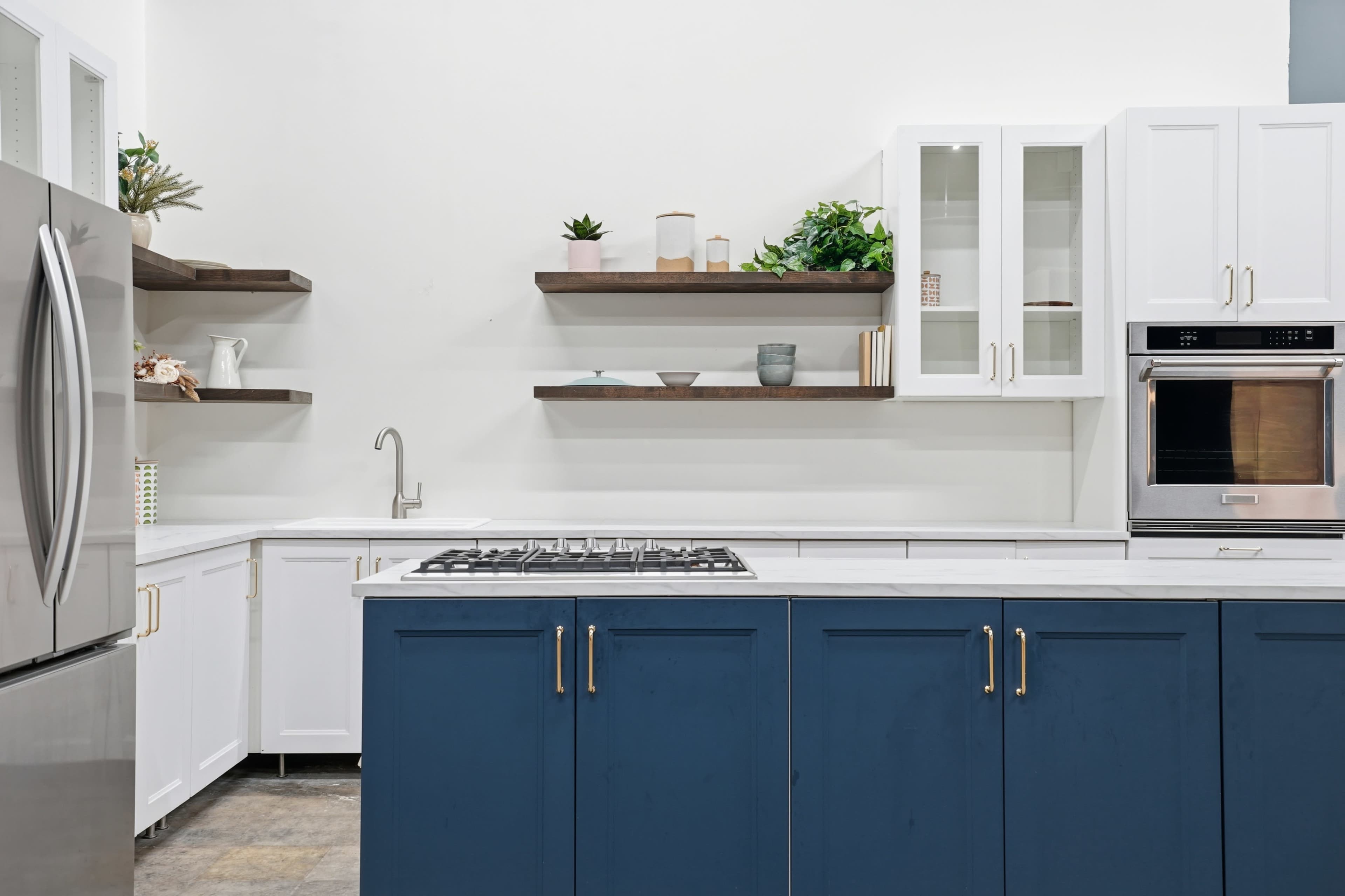 The kitchen features white cabinets with blue lower cabinets, a stainless steel refrigerator, a gas stove, and open shelving displaying plants and decorative items.