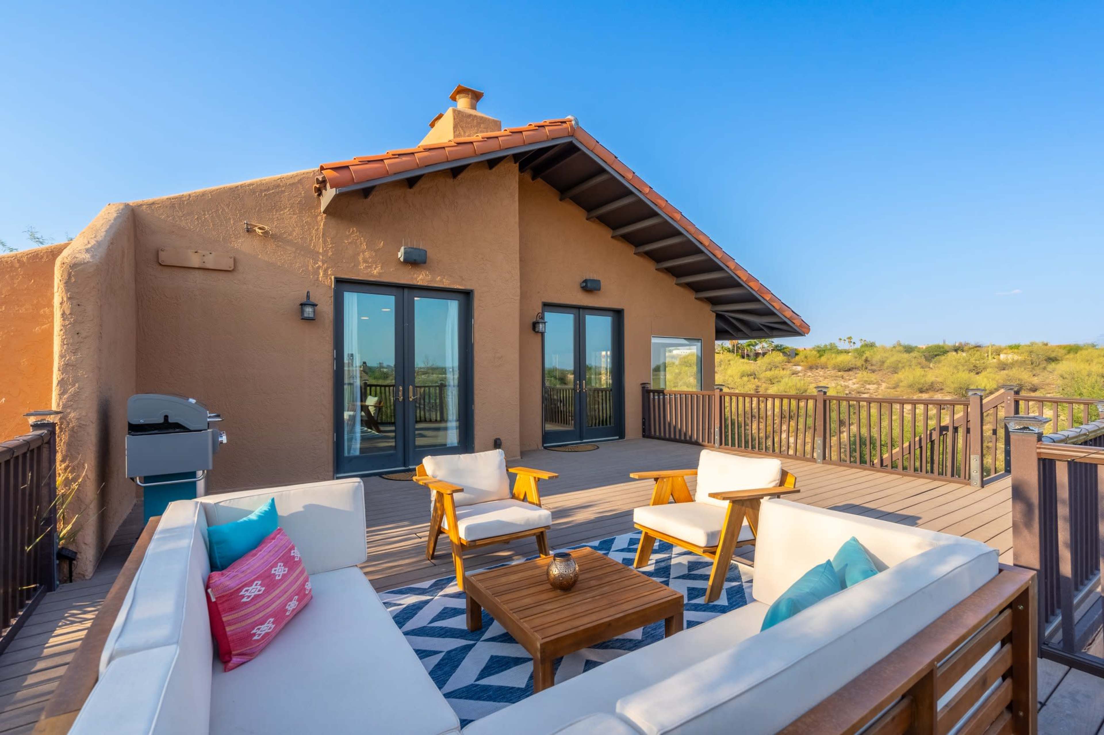 A spacious outdoor seating area features a modern furniture arrangement on a wooden deck, alongside a house with a sloped roof and sliding glass doors.