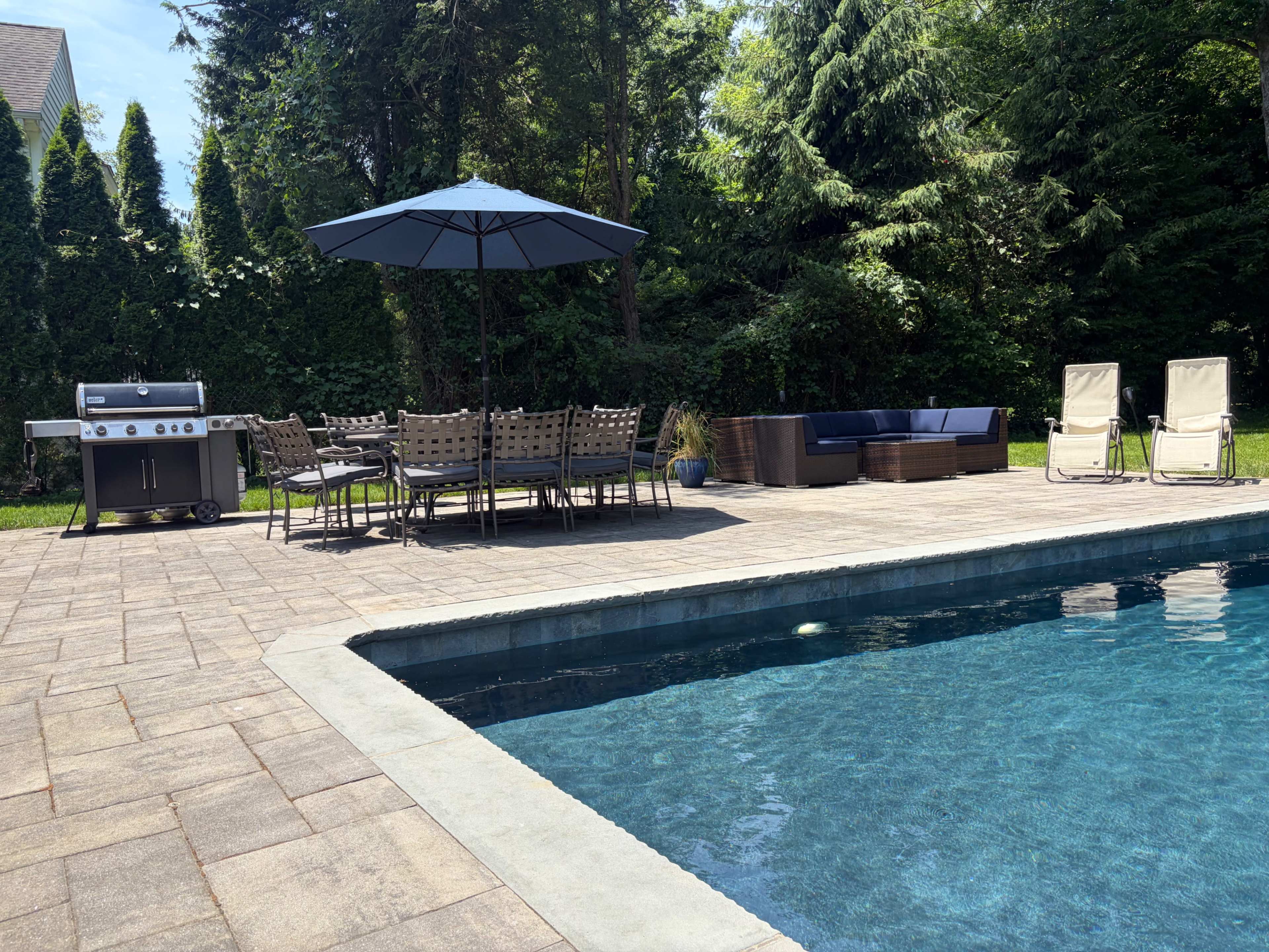 A pool area features a shaded dining set with a large umbrella, lounge chairs, a grill, and a sectional couch surrounded by greenery.