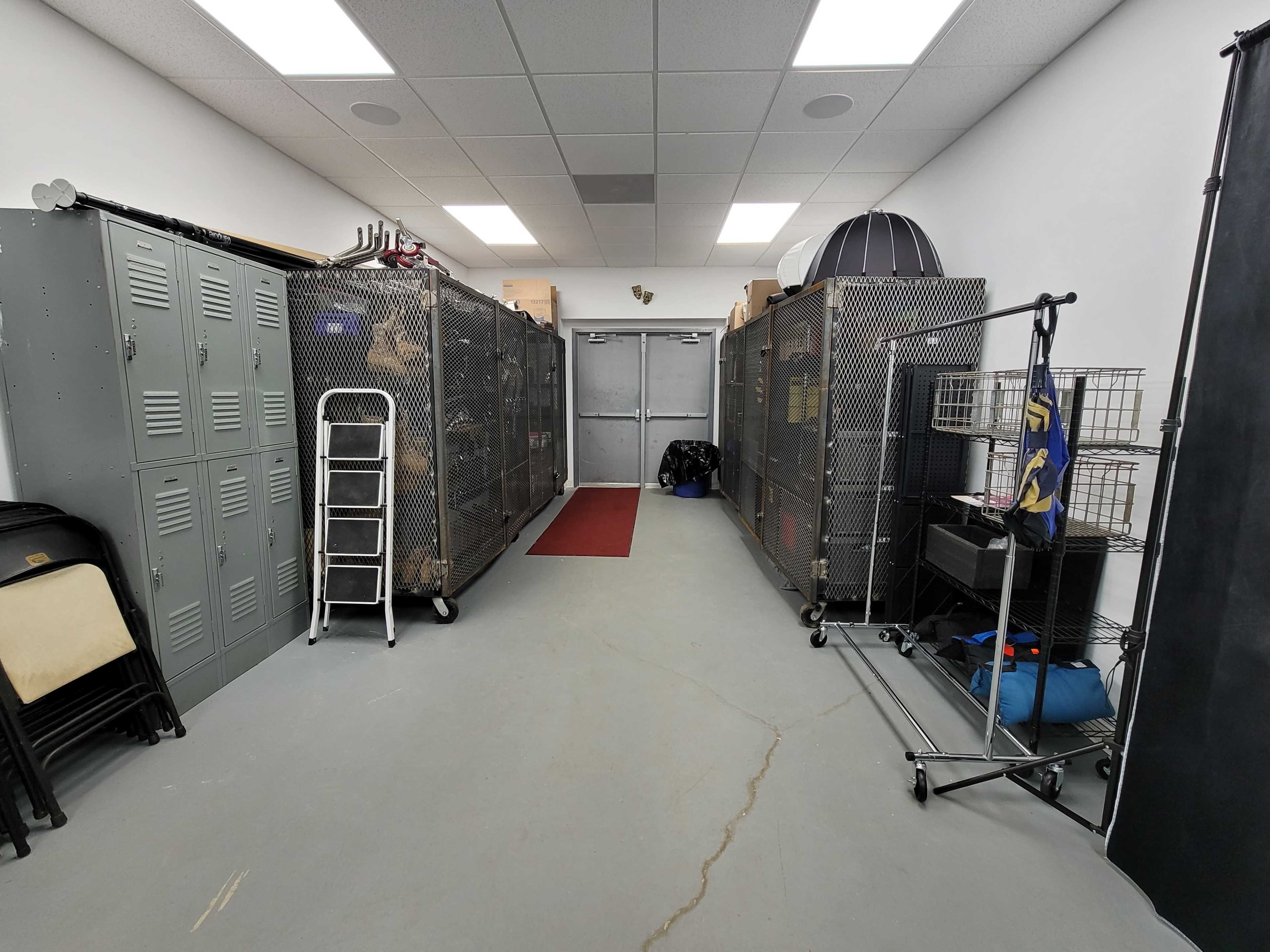 The image shows a hallway with metal lockers on one side and large wire cages on the other, leading to a double door at the end.