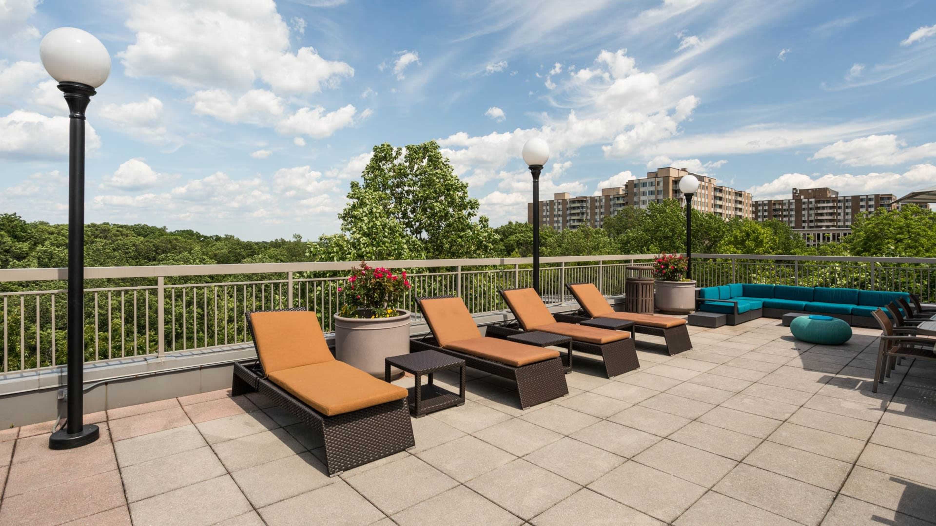 A rooftop patio features several lounge chairs and planters with flowers, overlooking a green landscape and modern buildings in the distance under a cloudy sky.