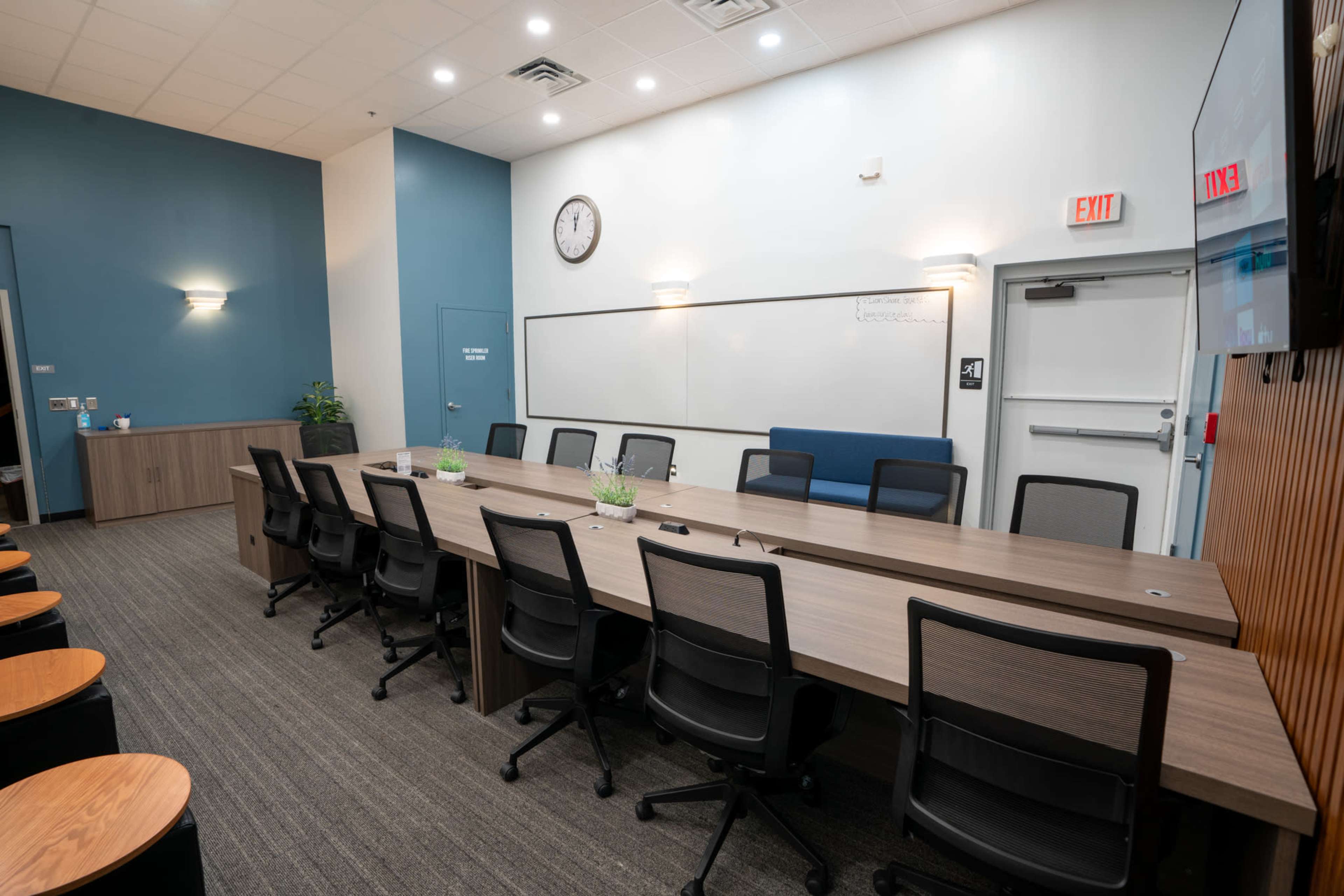 A conference room features a long, rectangular table with black chairs, a whiteboard, a clock on the wall, and a doorway leading to a restroom.