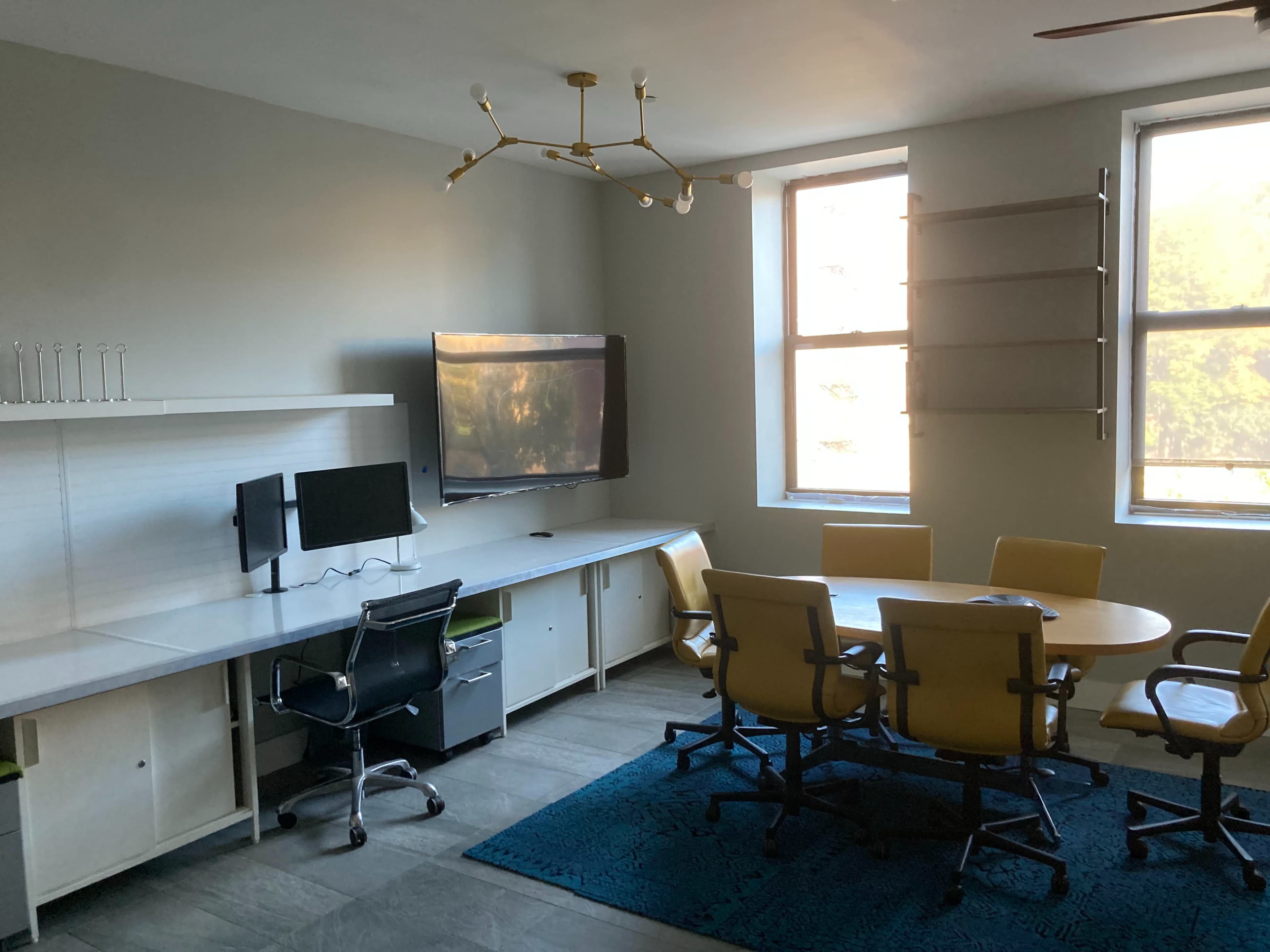 A modern office meeting room with a circular table surrounded by yellow chairs, a desktop computer setup, and a large screen mounted on the wall.