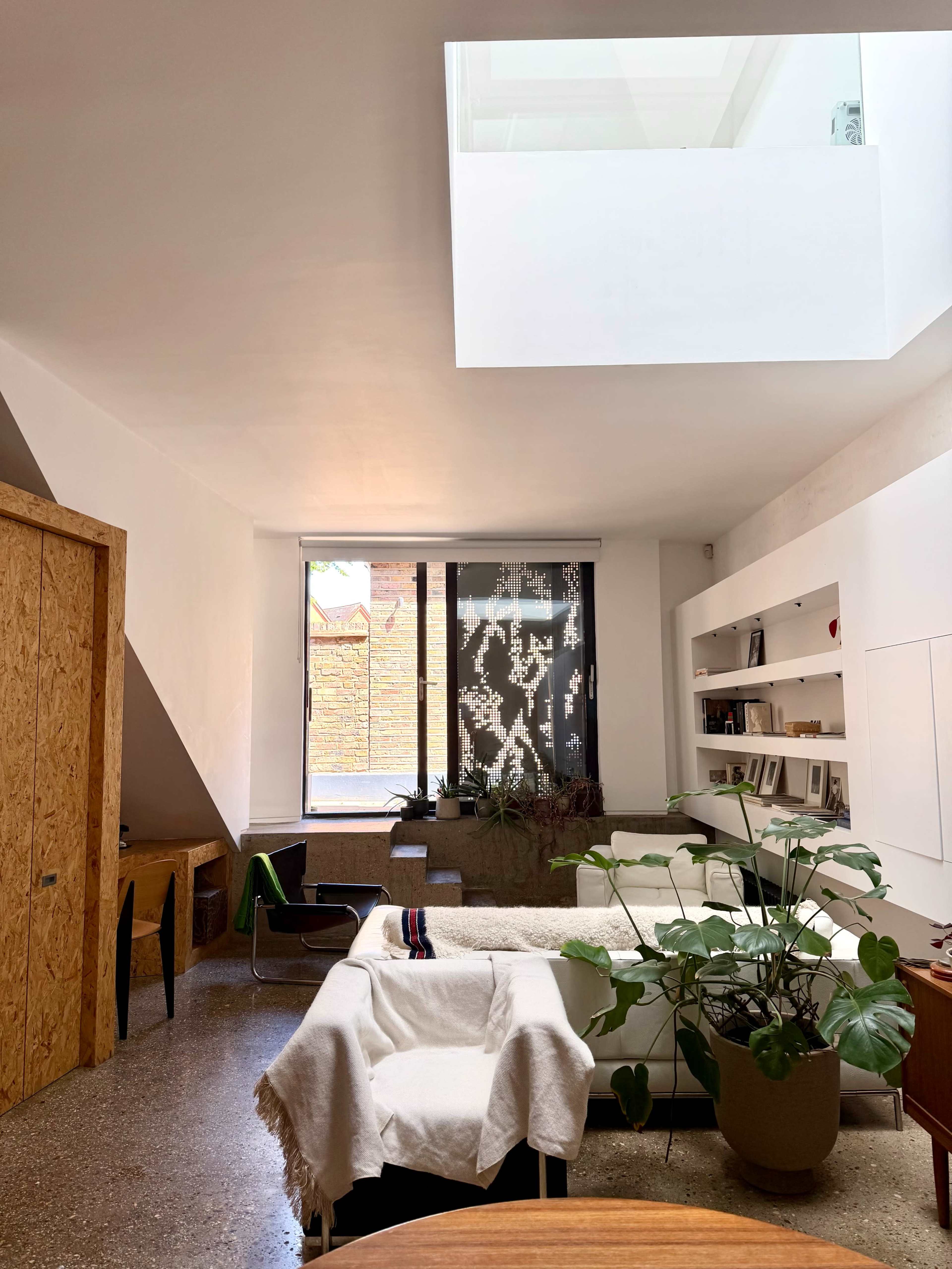 The image shows a modern living space featuring a white interior with a skylight, a large window, a seating area with a sofa, and a potted plant.