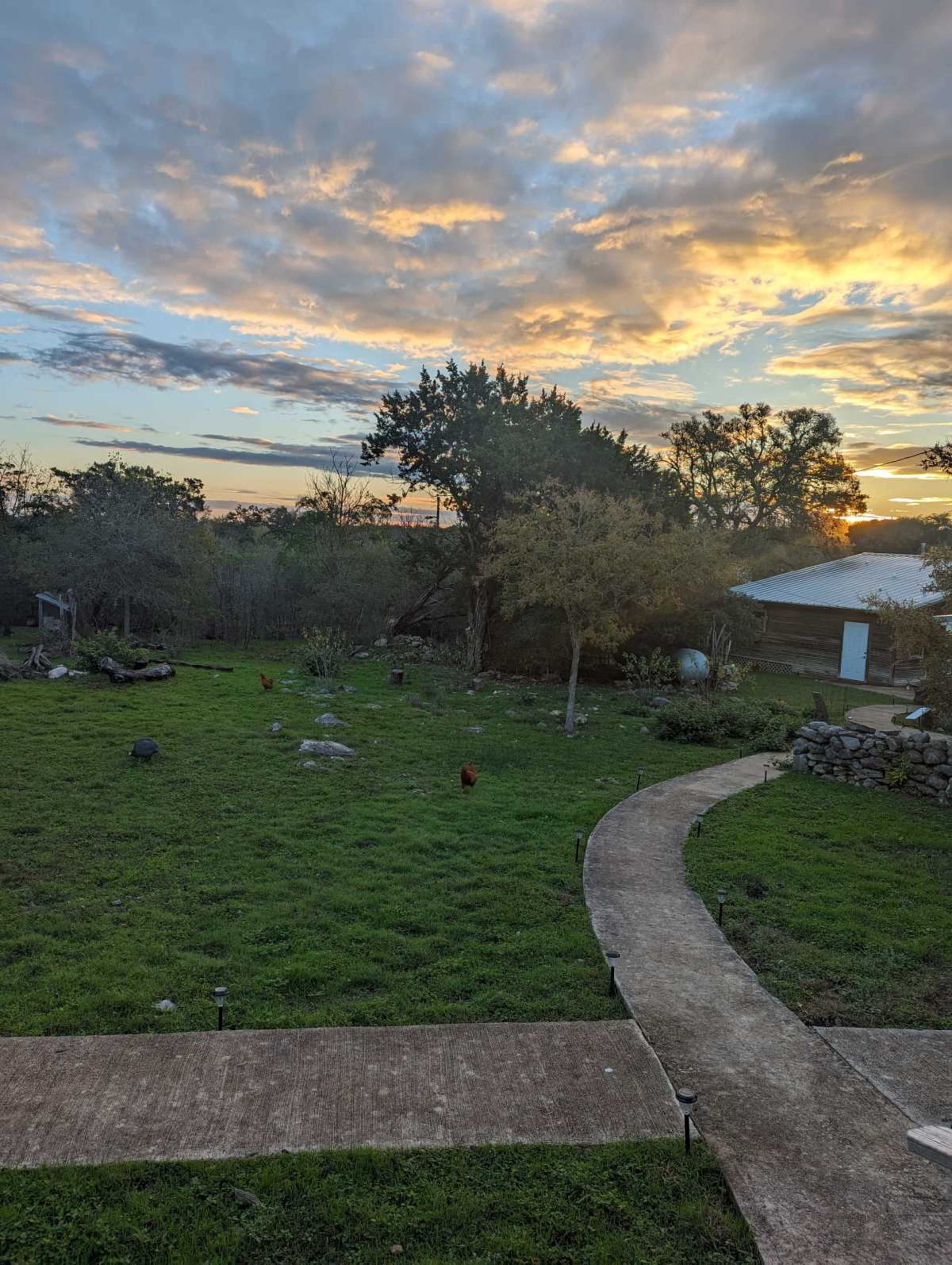 The image shows a grassy area with a winding path and a vibrant sunset in the background, featuring trees and a small building.
